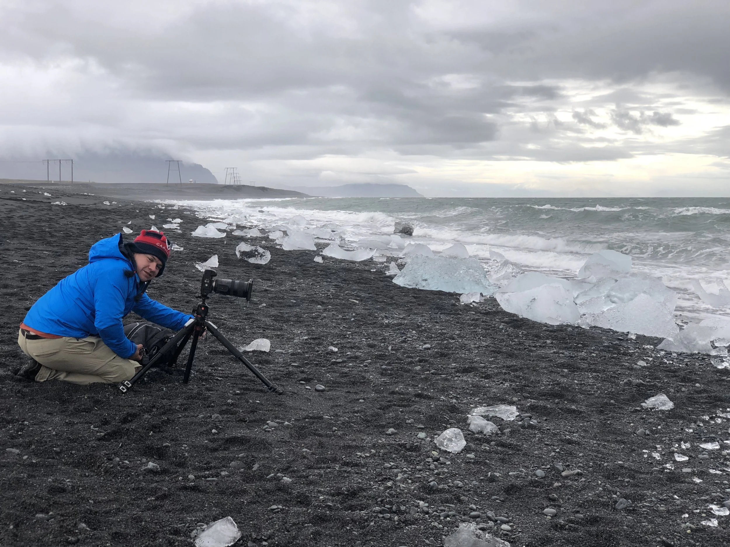 A man in a blue jacket crouches on a dark sandy beach with ice chunks scattered along the shoreline, holding a camera on a tripod, with ocean waves and a cloudy sky in the background.