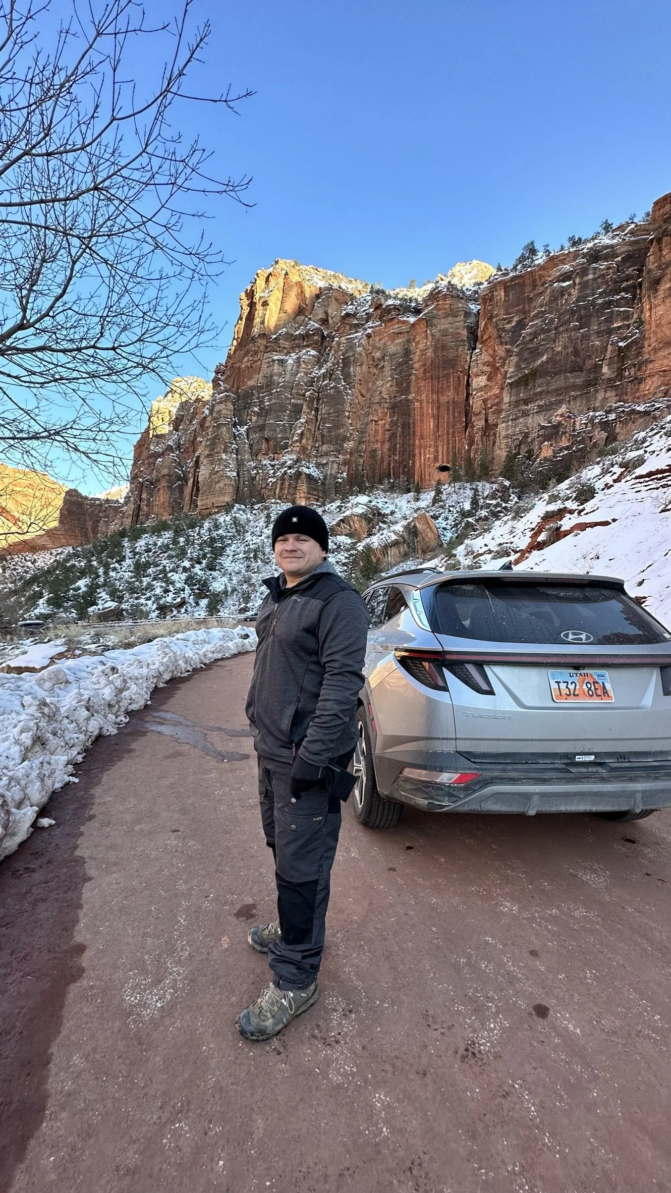 A man wearing a black beanie, black jacket, and hiking boots standing on a dirt road with snow on the sides. Behind him is a silver Hyundai Tucson, and towering red rock cliffs under a clear blue sky.