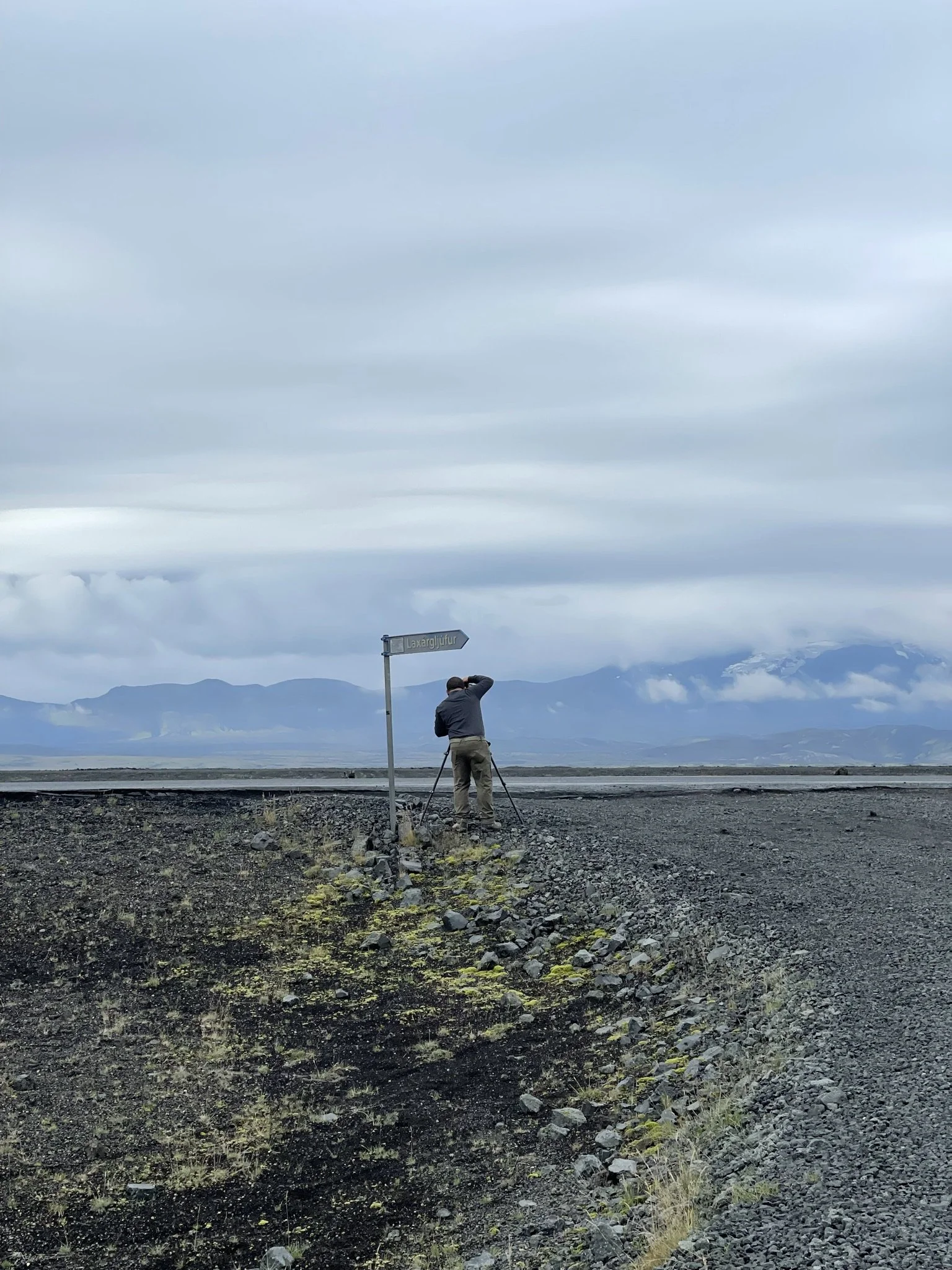 A person using a camera tripod stands on rocky terrain under a cloudy sky with mountains in the background.