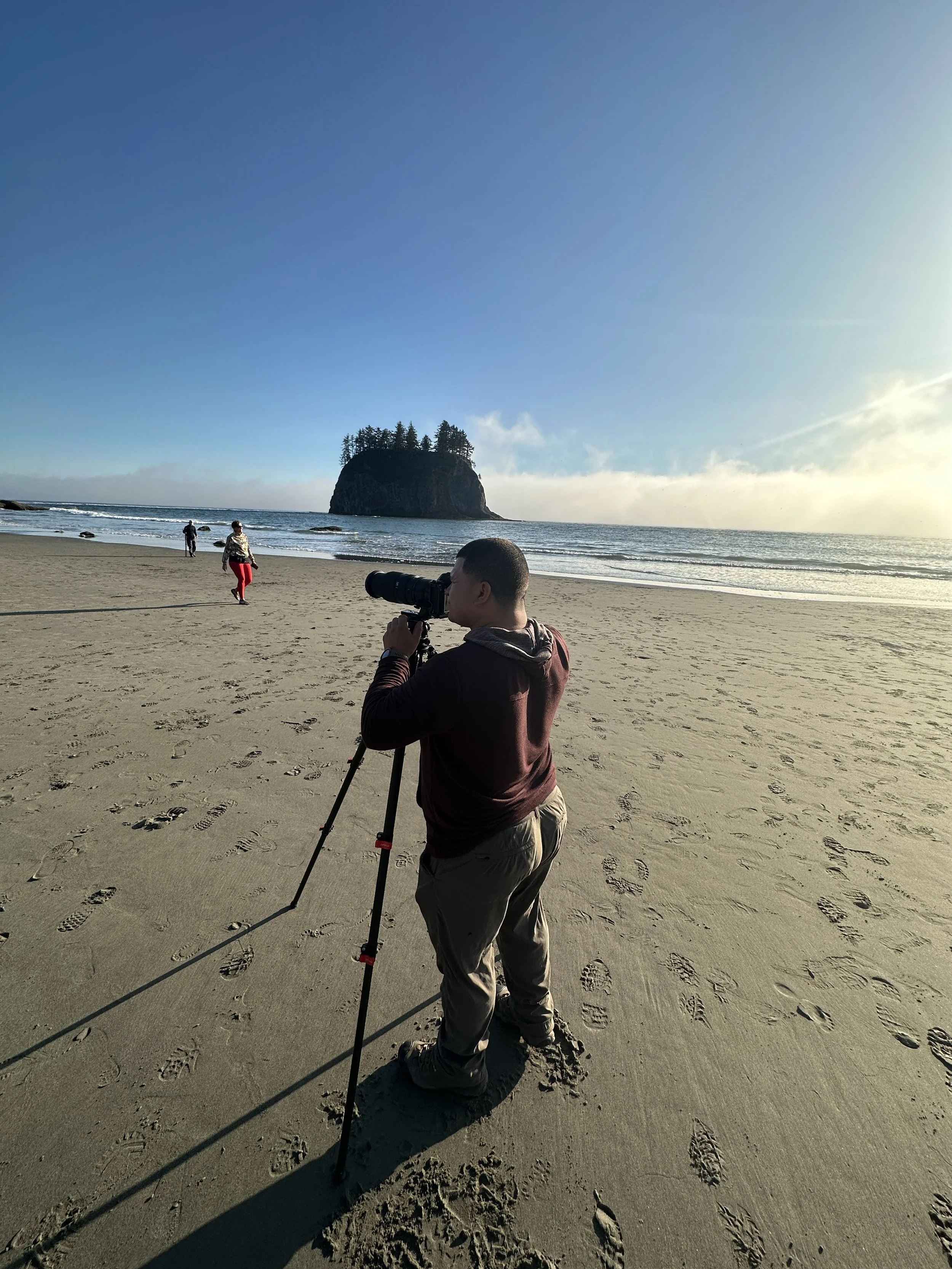 A person with a camera on a tripod taking photos on a sandy beach with footprints, ocean waves, a large rock formation with trees, and a clear sky in the background.