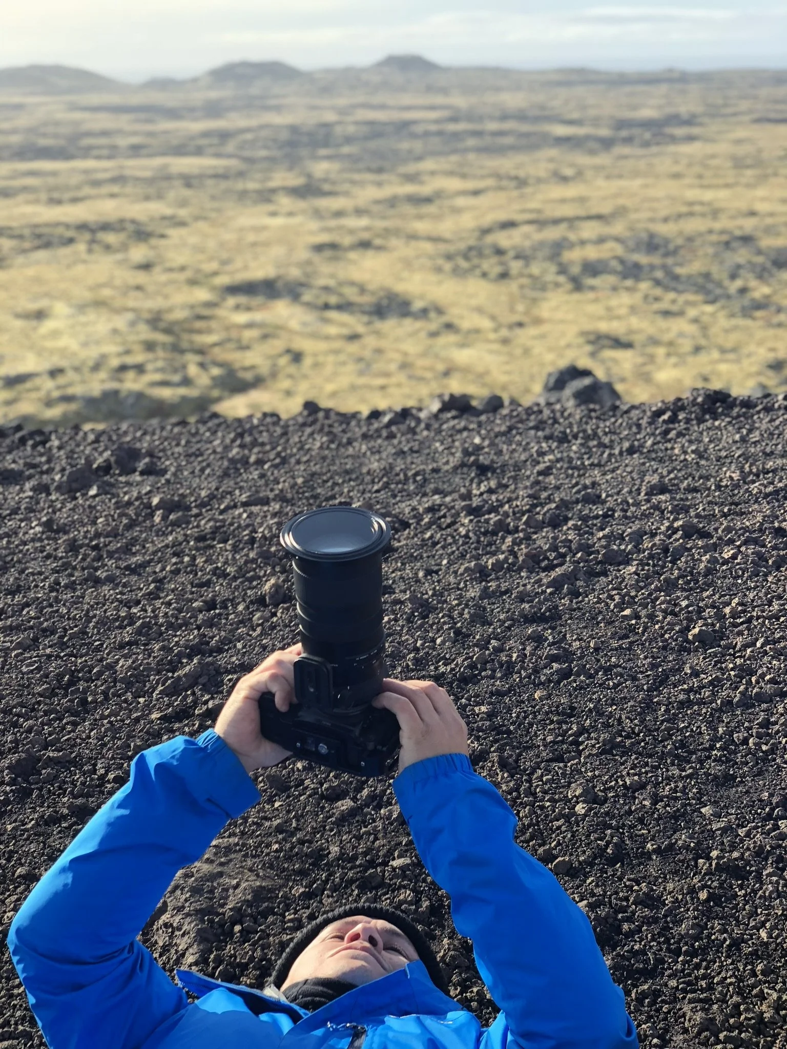 Person lying on dark volcanic terrain, wearing a blue jacket, taking a photograph with a camera pointed upward, with a vast, rocky landscape in the background.