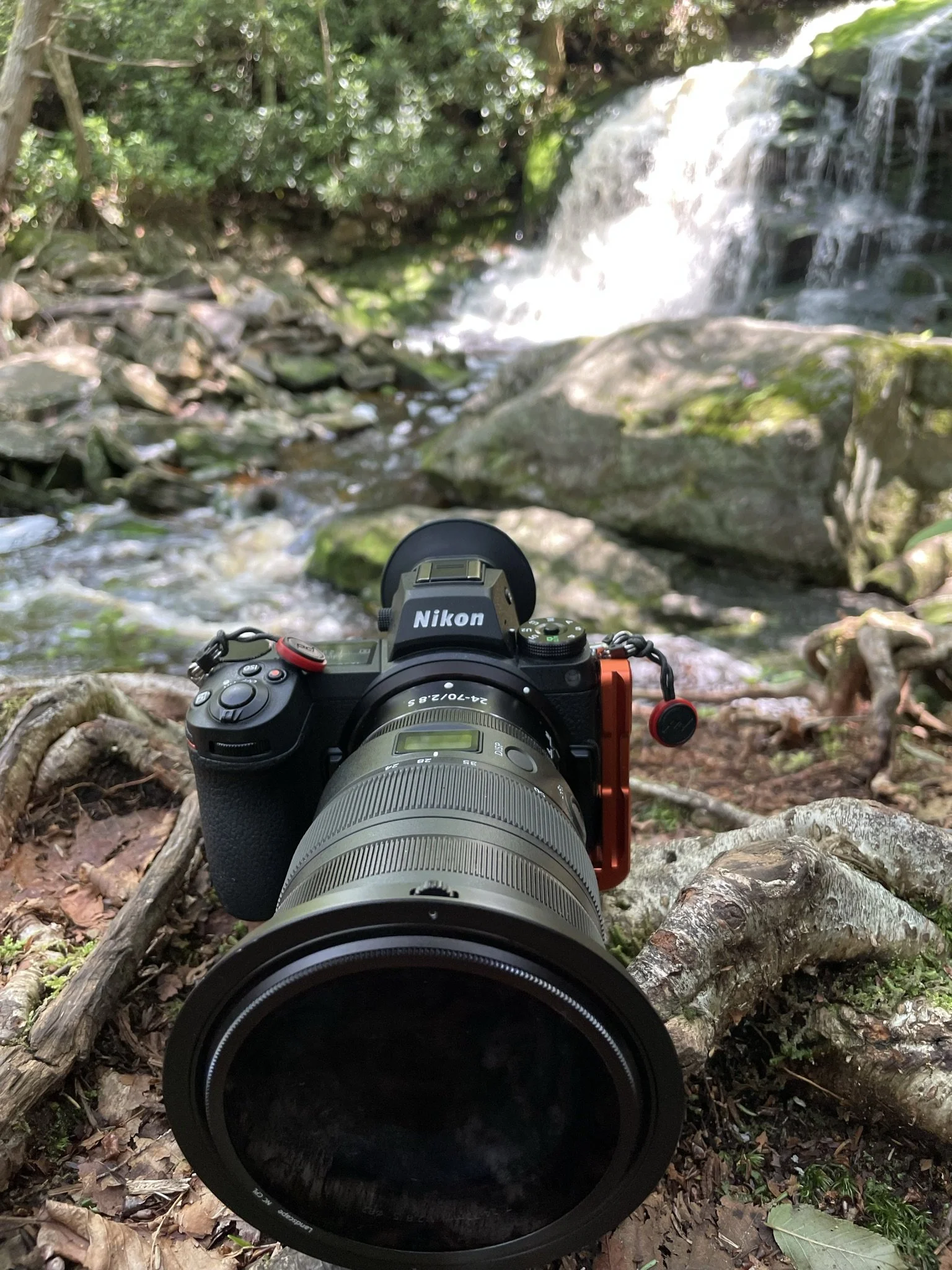 A Nikon camera with a telephoto lens is set up outdoors on a tree root, aiming toward a waterfall cascading over rocks in a lush green forest.