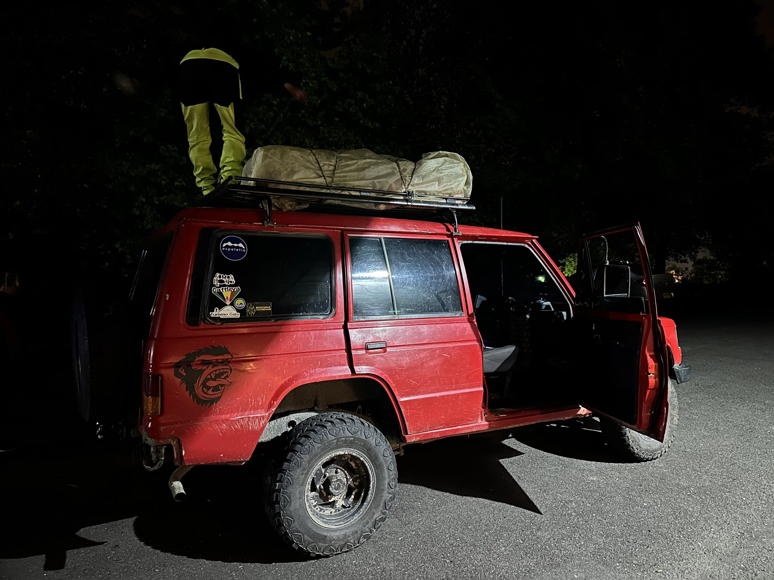 A red off-road vehicle with stickers and a lion face decal on the side, parked at night. The driver's door is open, and there is a person standing on the roof rack, which is loaded with a large covered item. The vehicle is on a paved surface near tre