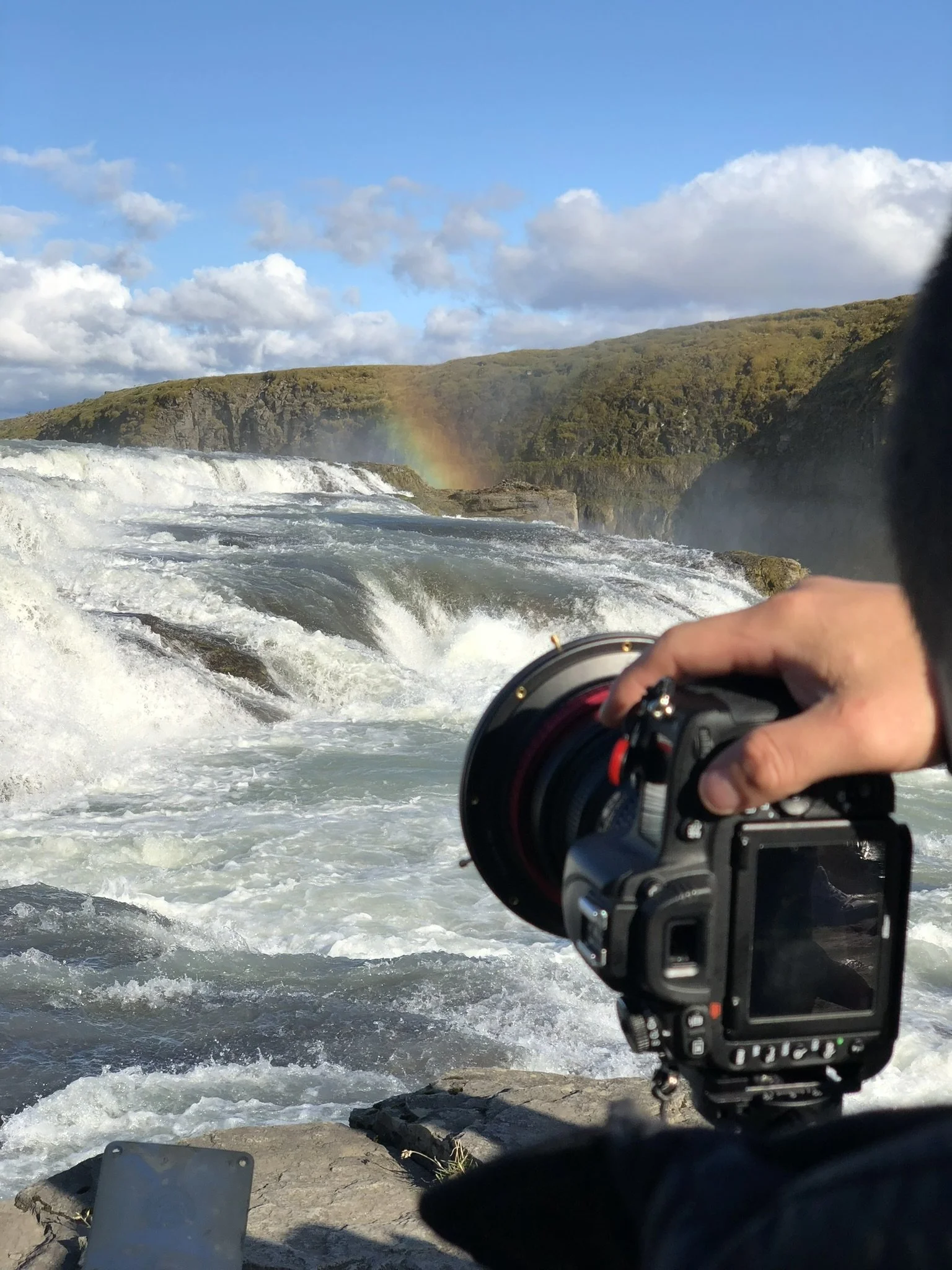 Photograph of a person holding a camera near a river with rushing water, green hills, and a rainbow in the sky in the background.
