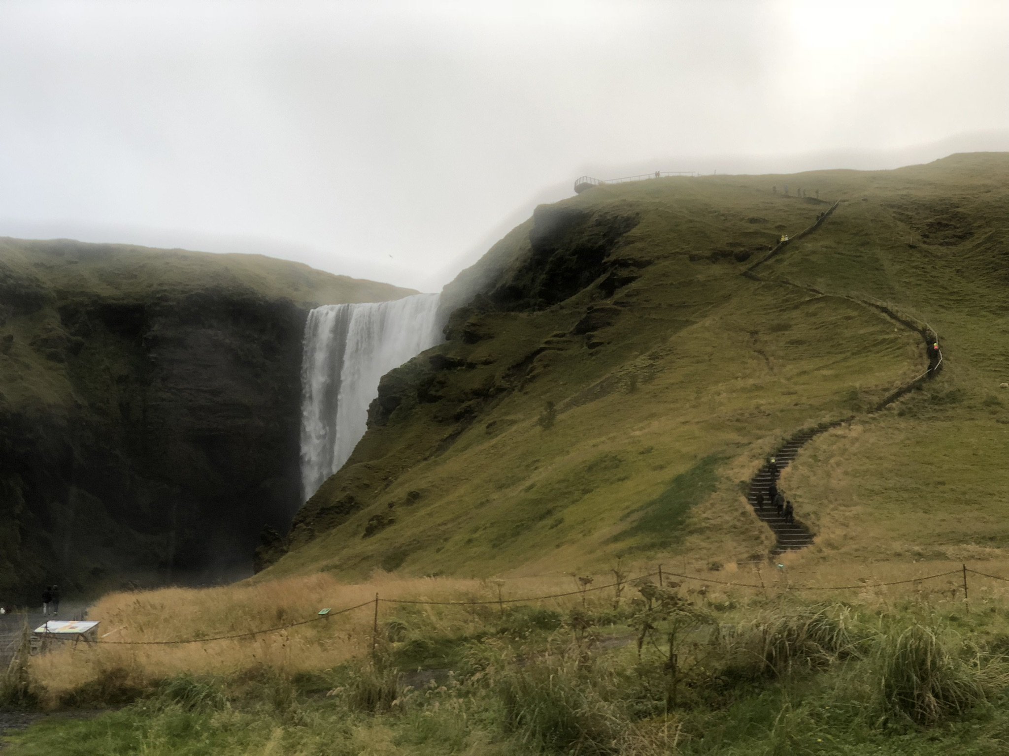 A waterfall flowing down a steep cliff surrounded by green grassy hills, with a winding staircase and pathway leading up the hill, and mist in the overcast sky.