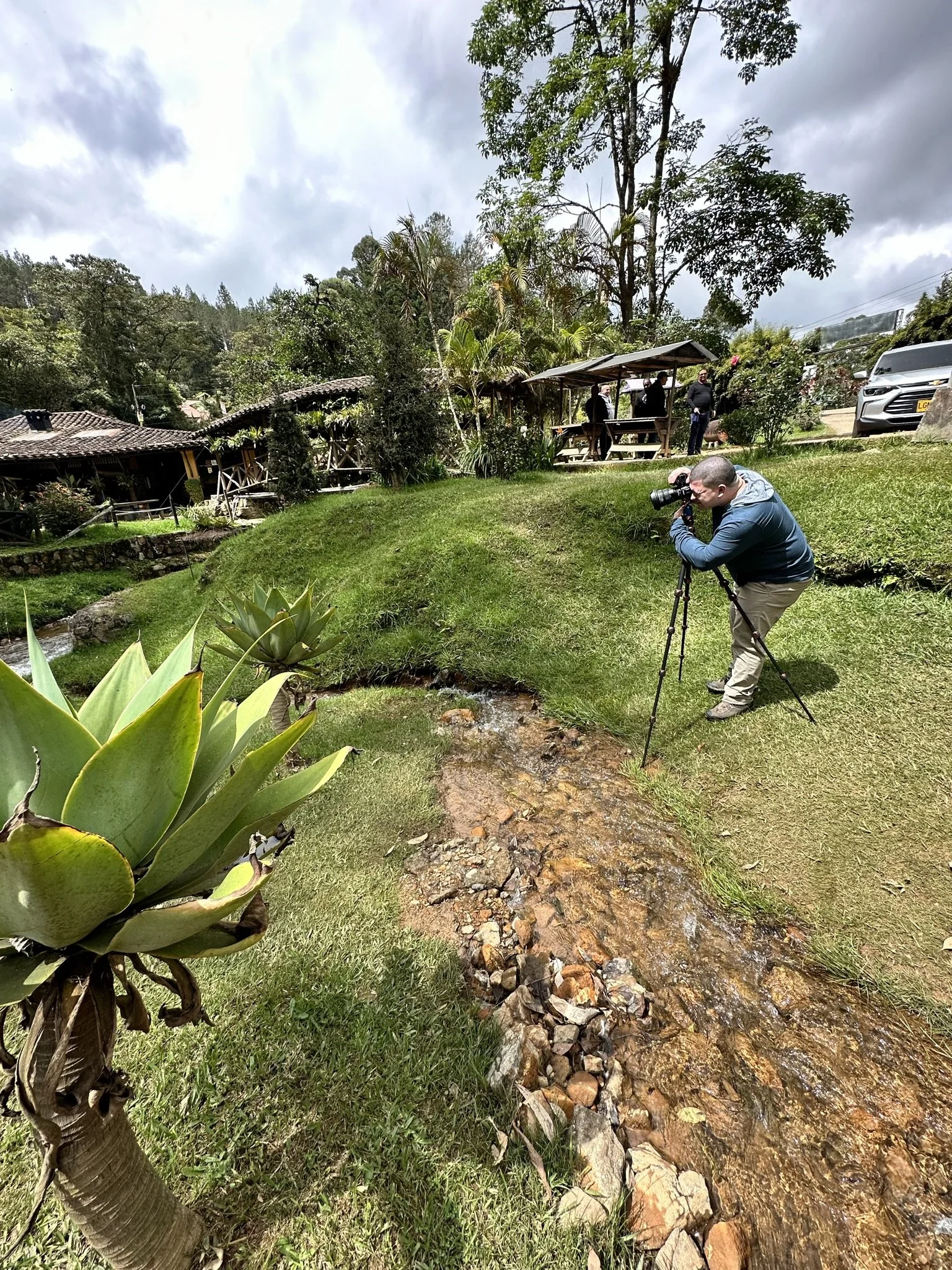 A man taking a photograph of a small stream in a lush green outdoor setting with trees, bushes, and wooden structures under a cloudy sky.