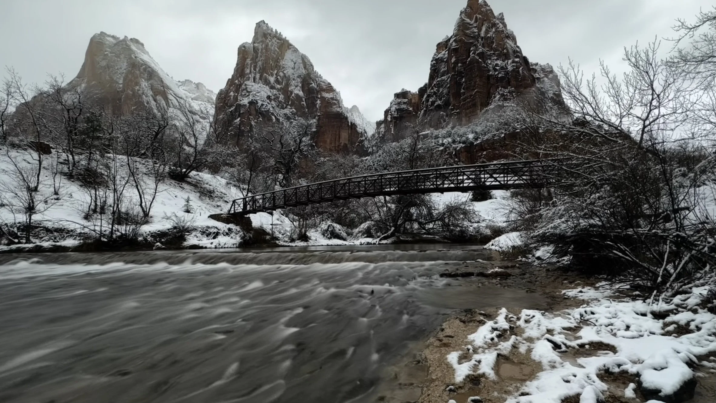Snow-covered rocky mountains with a small wooden bridge over a flowing river in a winter landscape.