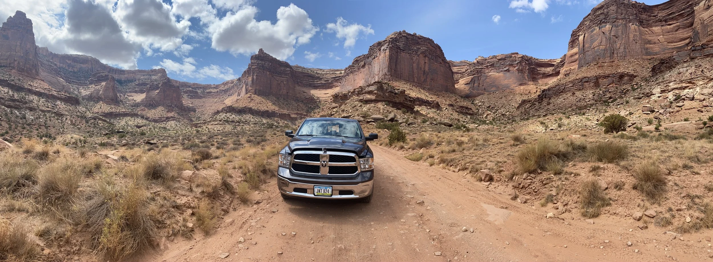 A black pickup truck driving on a dirt trail in a desert landscape with large red rock formations and cliffs in the background under a partly cloudy sky.
