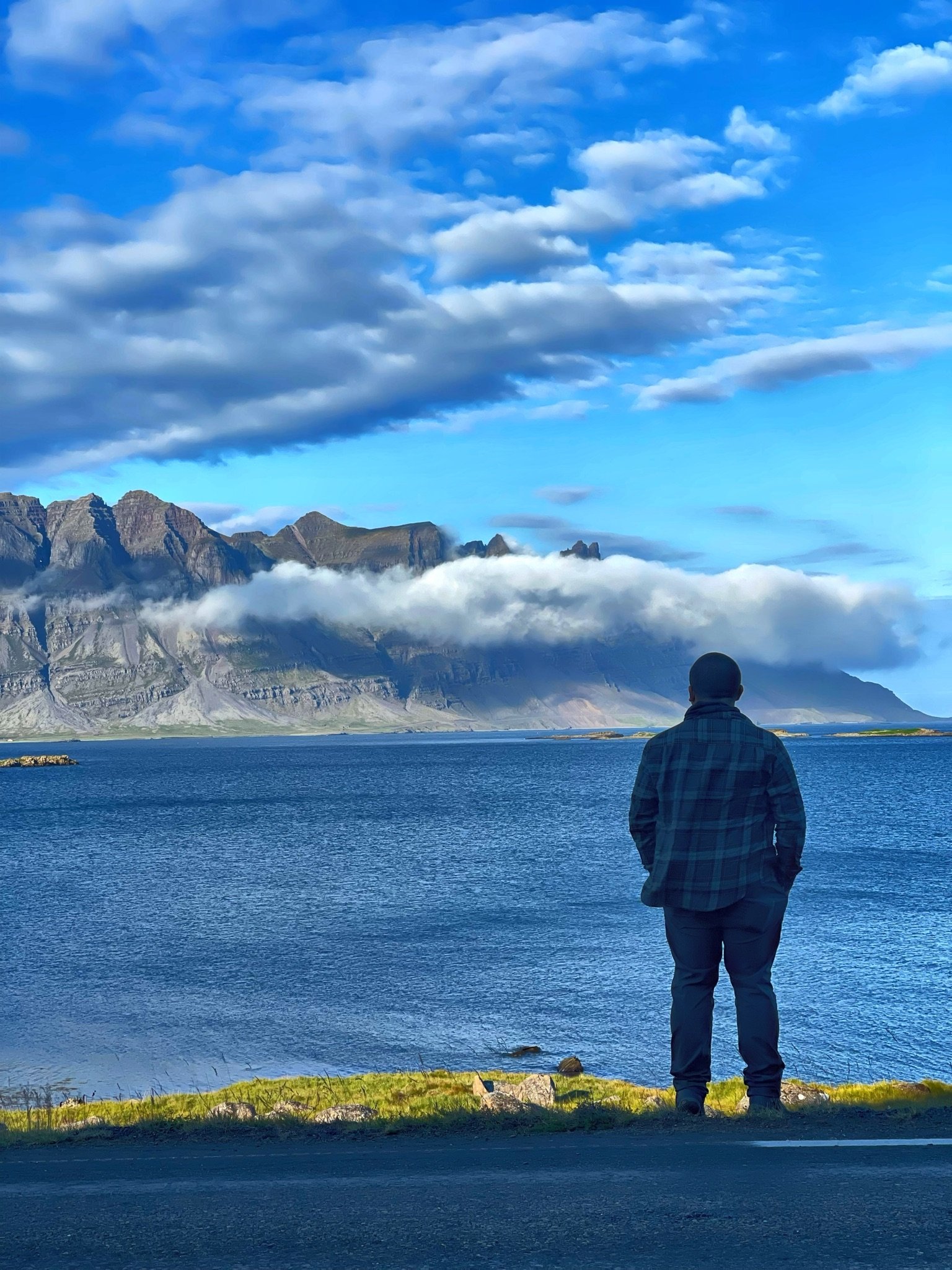 A person stands on the edge of a shoreline, looking out at a mountain range with low-hanging clouds over the mountains, a body of water, and a sky filled with clouds.