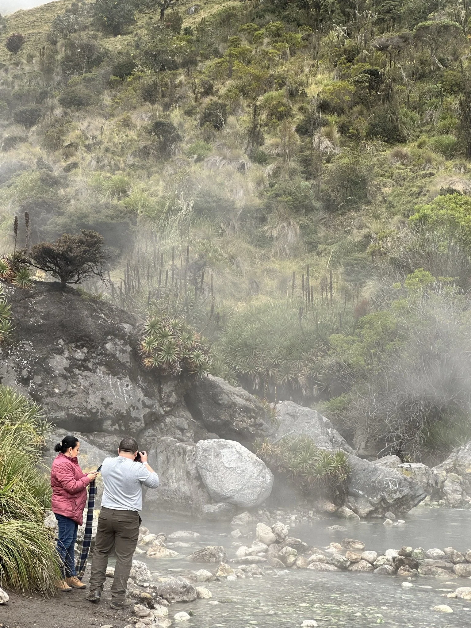 Two people standing near a rocky riverbank in a lush, foggy, mountainous forest, with one person taking a photo and the other looking at their phone.