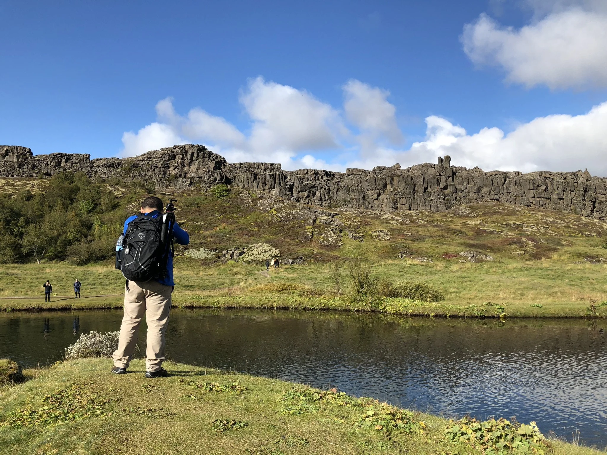 Hiker with backpack looking at a small pond on a grassy landscape, with rocky cliffs and a partly cloudy blue sky in the background.