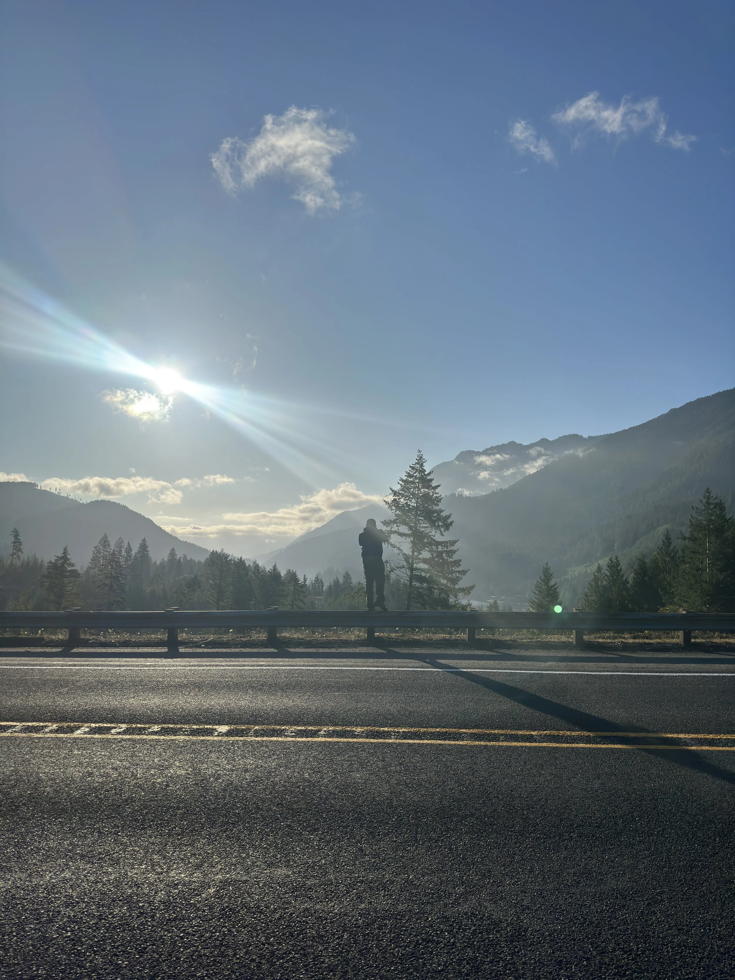 Person standing on the side of a mountain road, looking at the scenery with mountains, trees, and clouds in the background, under a bright sun in a clear sky.