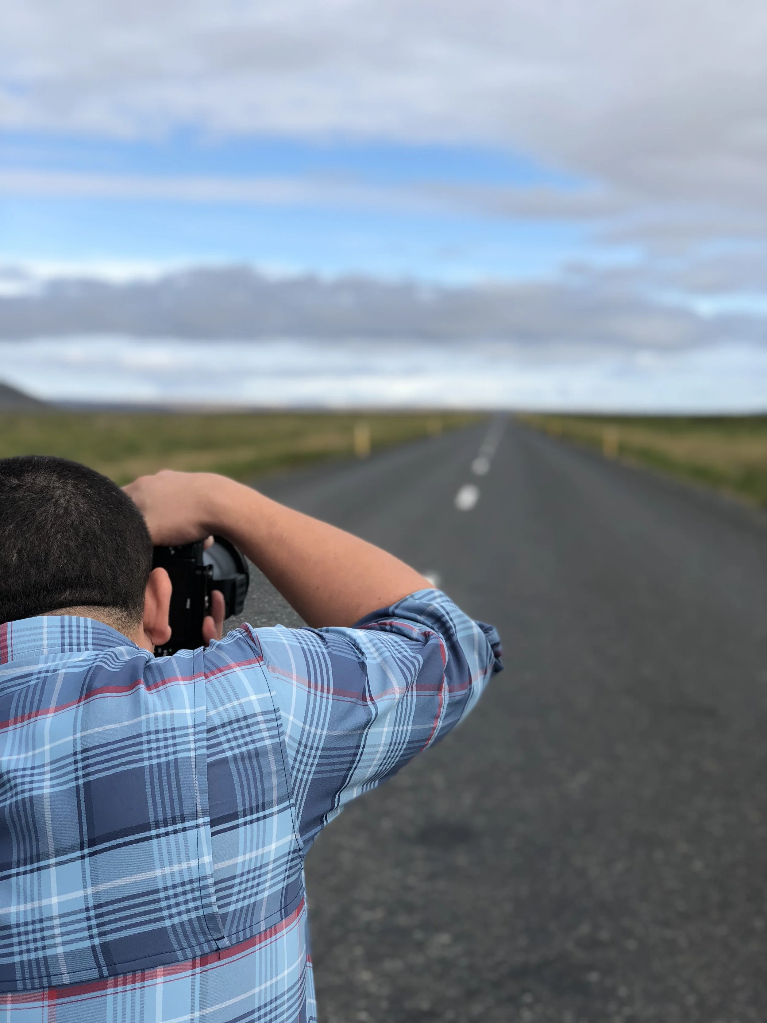 A person wearing a blue plaid shirt taking a photograph of an empty two-lane road that stretches into the horizon with green fields on either side under a partly cloudy sky.