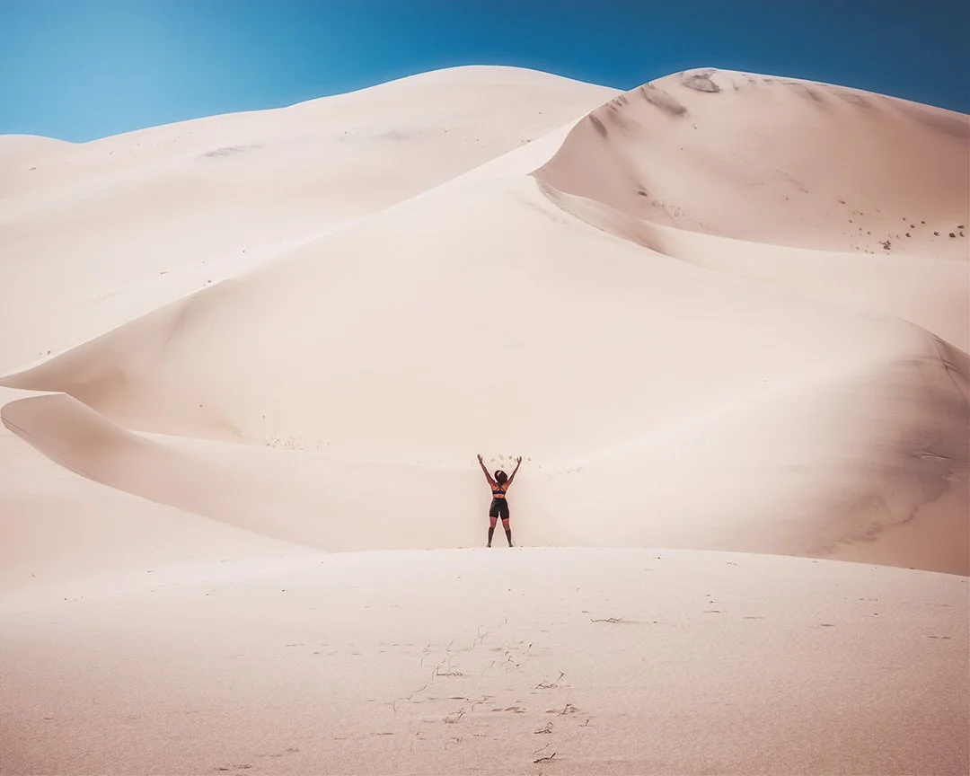 A person standing in a vast desert of white sand dunes, with arms raised, under a clear blue sky.