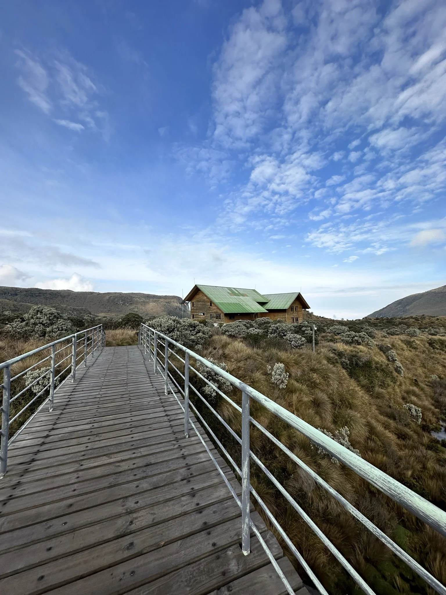 A wooden walkway with metal railings leading towards a cabin on a grassy hillside under a partly cloudy blue sky.