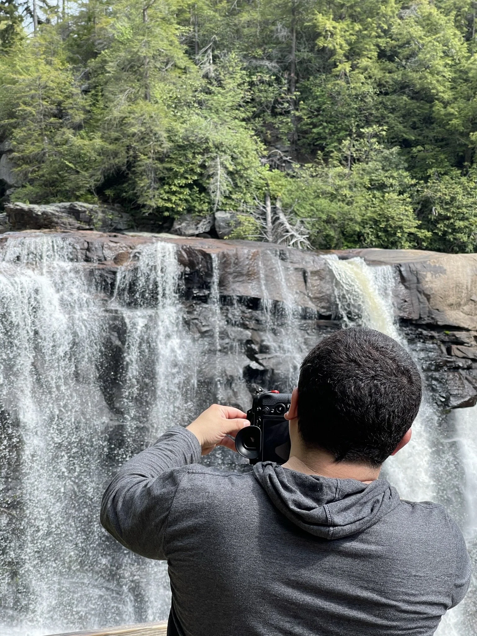 A man with short dark hair, wearing a gray hoodie, is taking a photo of a waterfall in a forest with a camera, viewed from behind.
