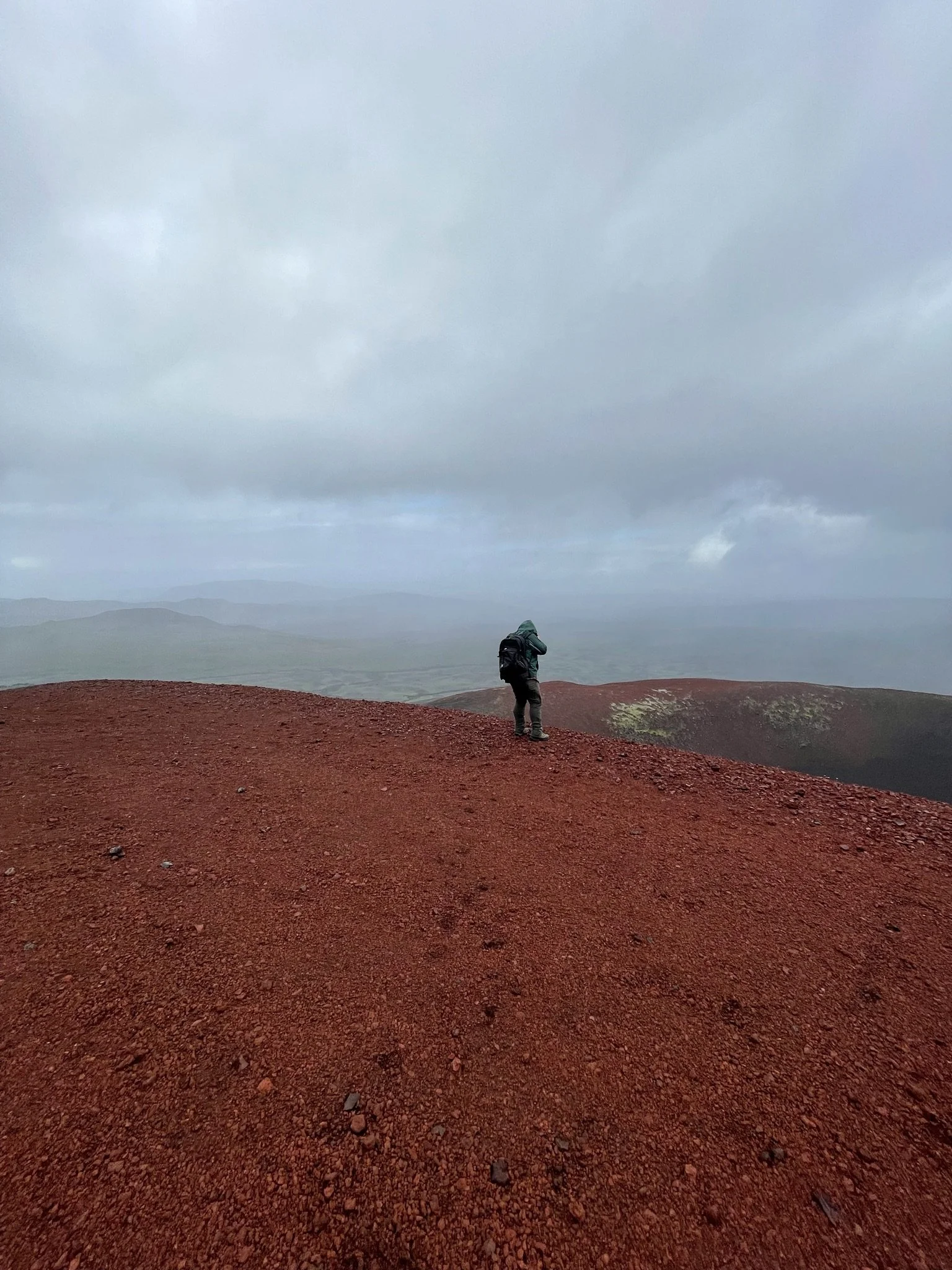 A person with a backpack standing on a reddish-brown, rocky hilltop on a cloudy day, with distant hills and a foggy sky in the background.