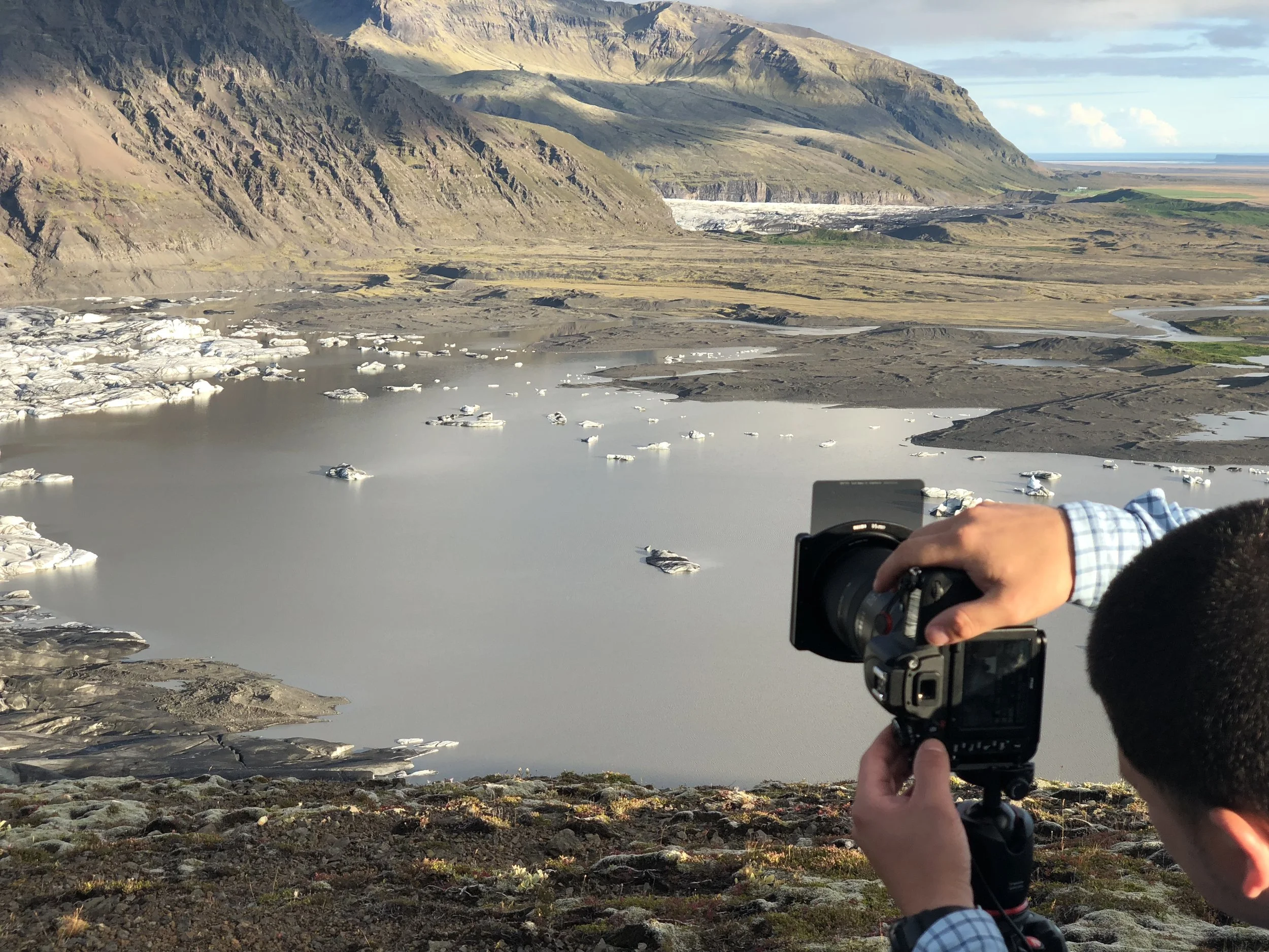 A person is taking a photograph of a landscape with floating icebergs on a body of water, surrounded by mountains and a valley.