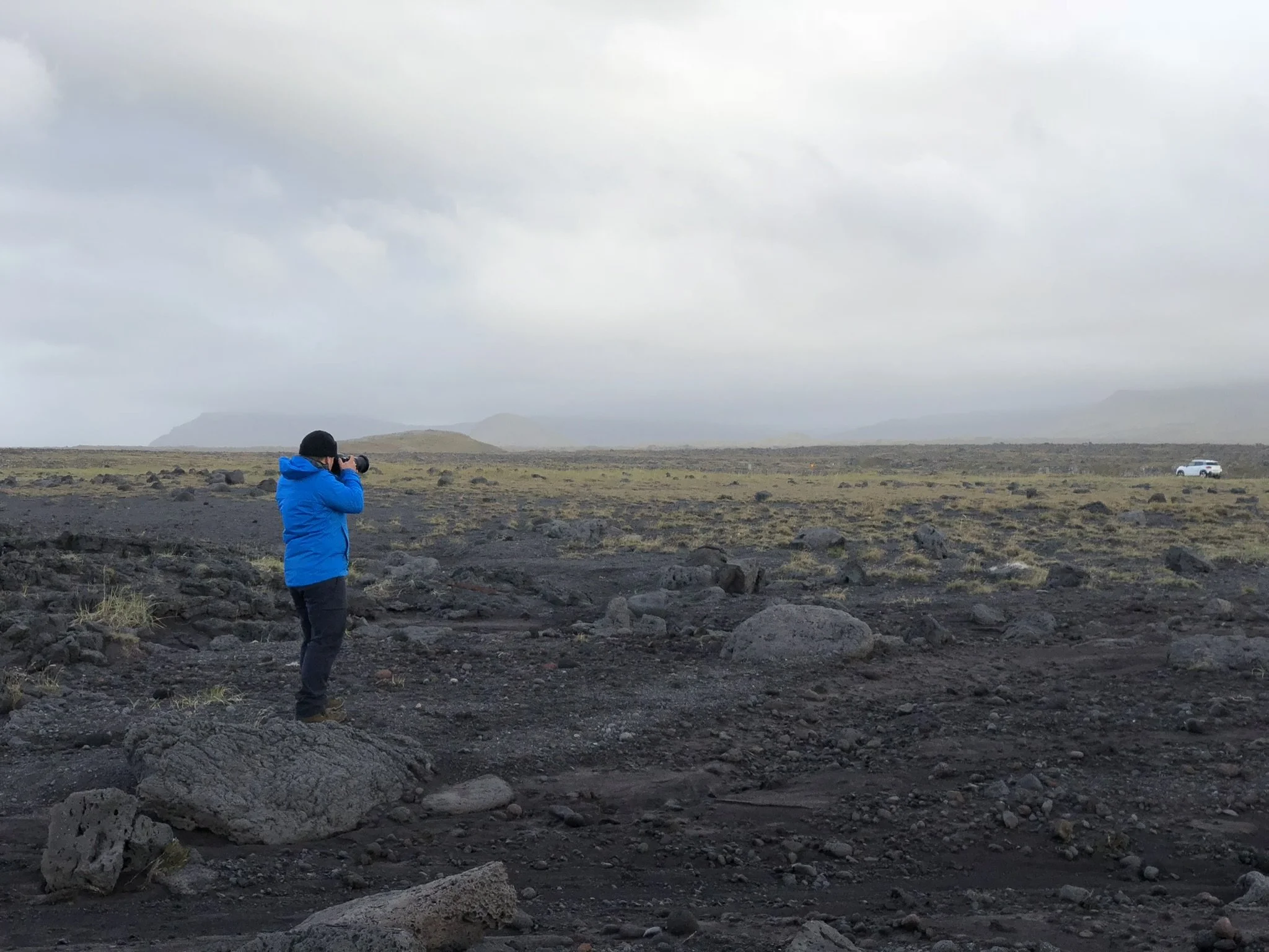 Person in a bright blue jacket taking a photograph in a rocky, volcanic landscape with mountains in the distance and a white vehicle parked far away.