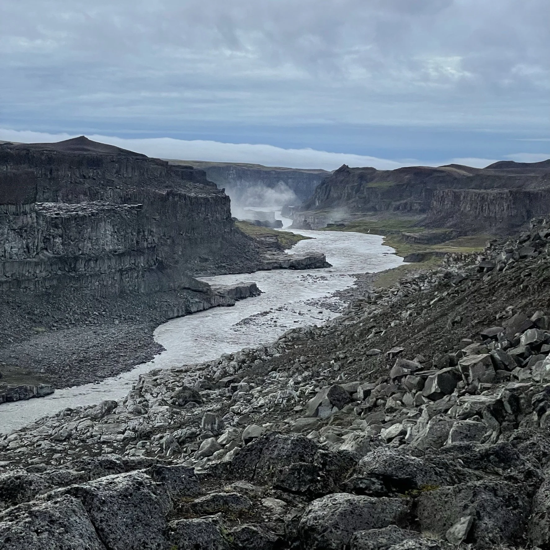 A rugged river flowing through a steep canyon with rocky cliffs and gravel on the slopes, under a cloudy sky with mist rising from the water.