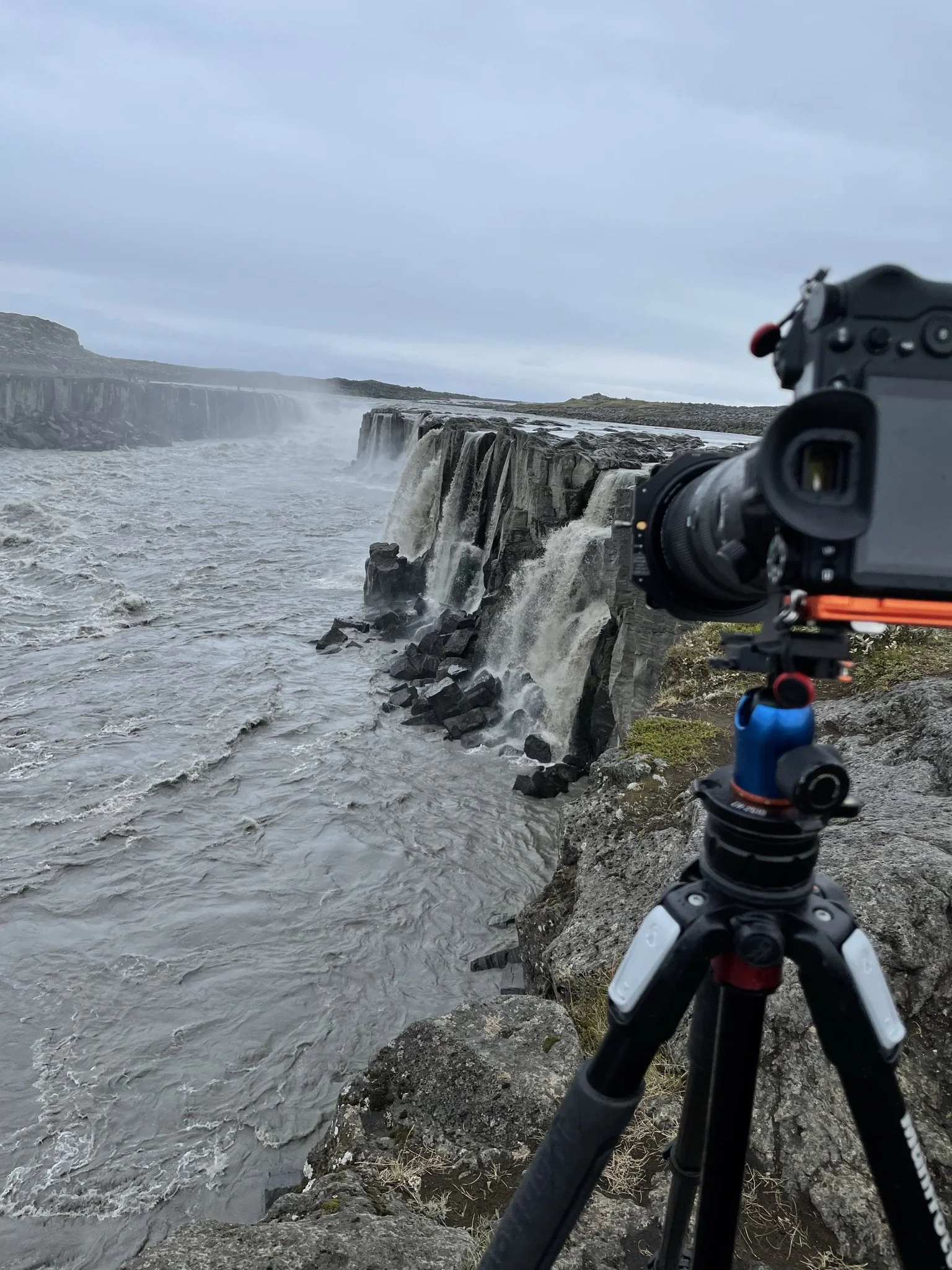 A camera on a tripod positioned near a waterfall with cascading water in a rugged outdoor setting under cloudy skies.
