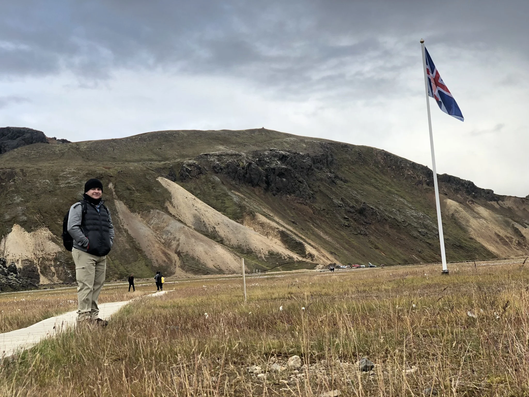 A person stands on a grassy path in front of a large flagpole with the Union Jack flag, in a landscape with rolling hills and overcast sky.