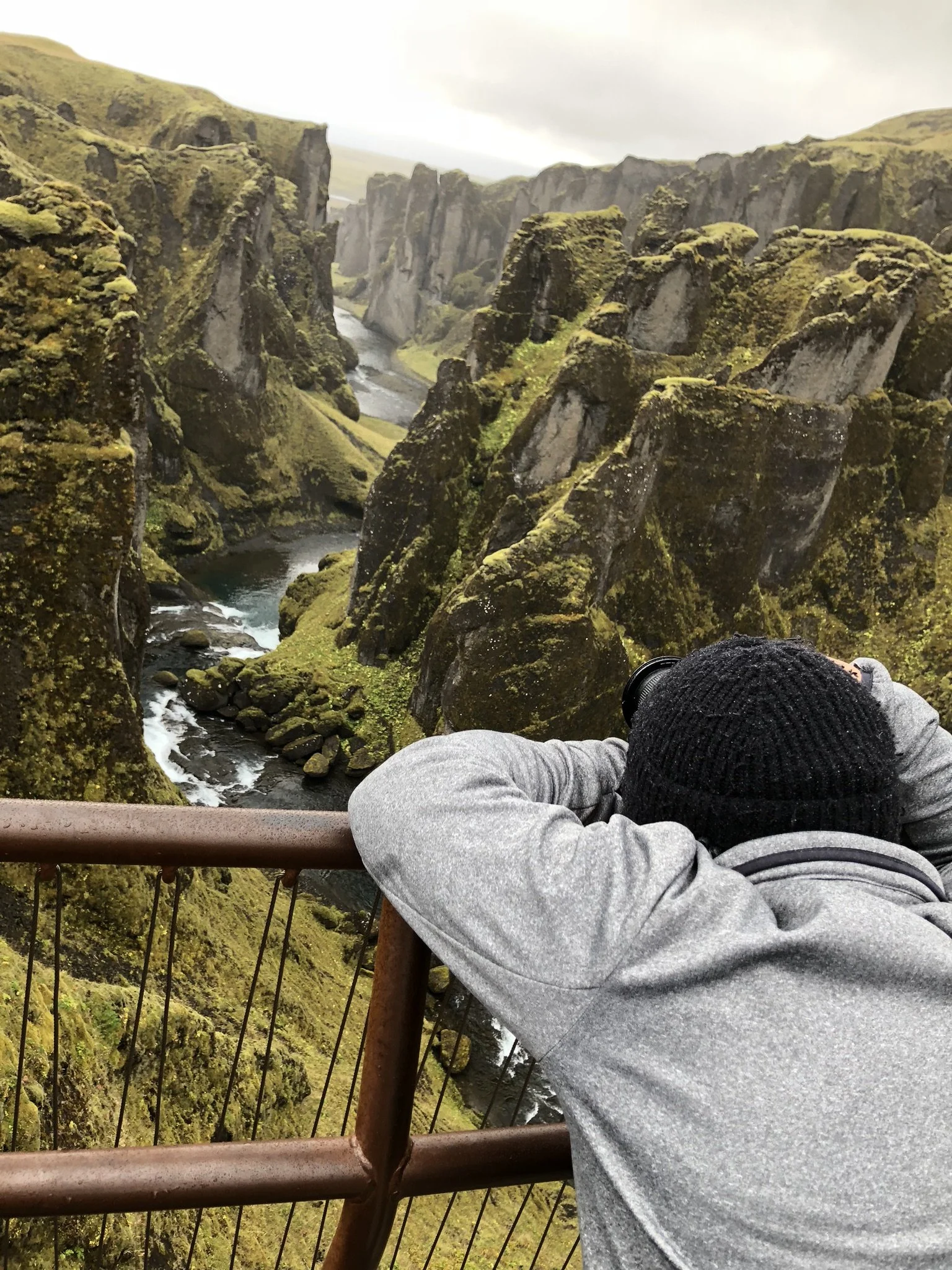 Person in gray hoodie and black knit hat taking a photo of a moss-covered canyon with waterfalls and a river, from a viewing platform with a rust-colored railing.