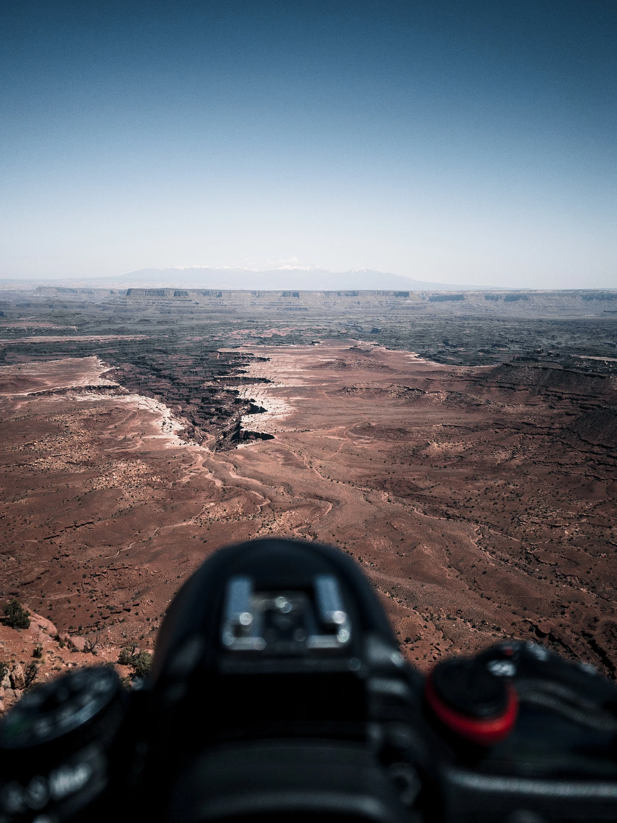 Looking down from a mountain with a drone controller in the foreground, vast desert landscape with canyons and a distant mountain range under a clear blue sky.