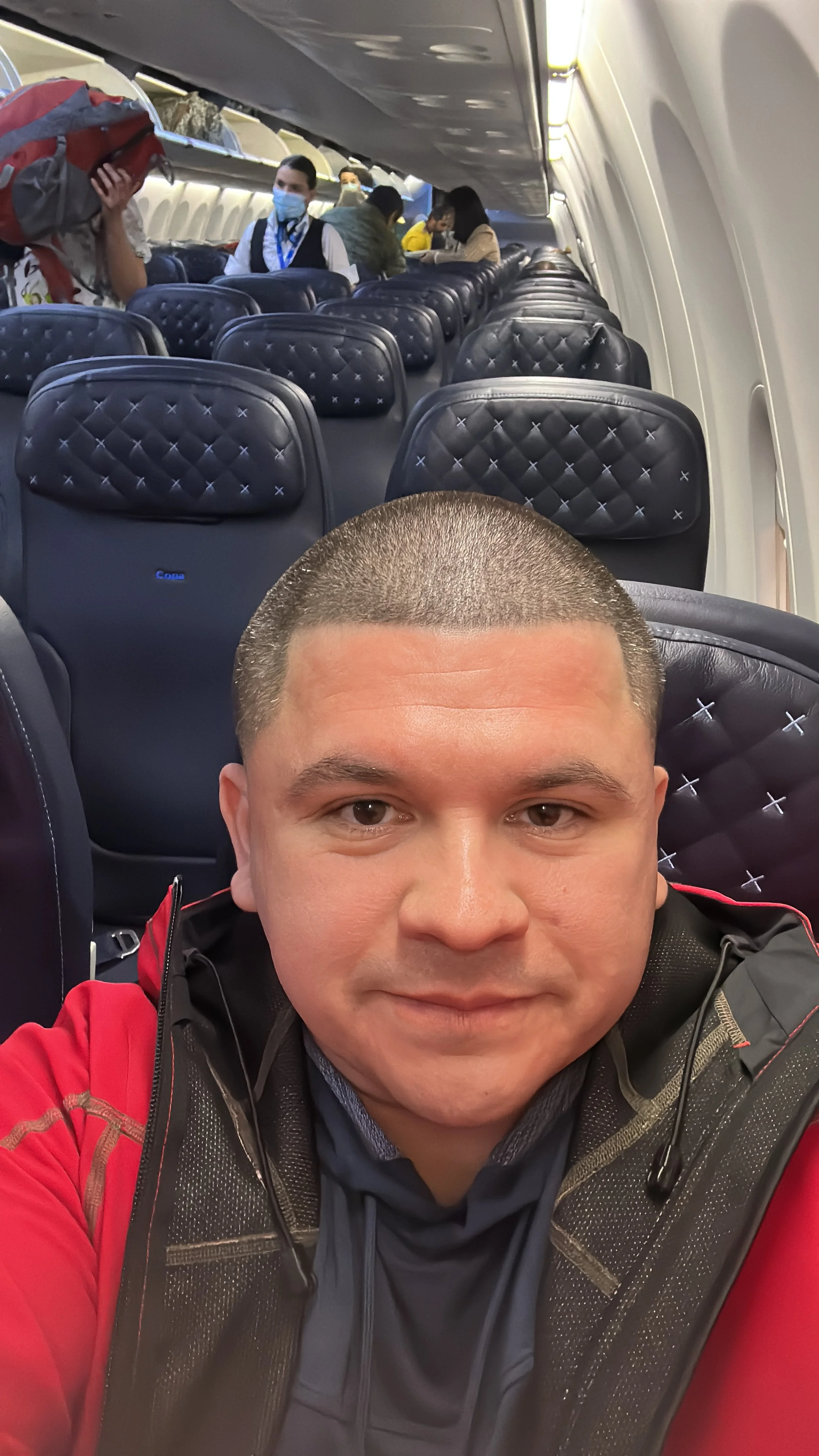 A man taking a selfie inside an airplane cabin with empty seats behind him and other passengers at the back.
