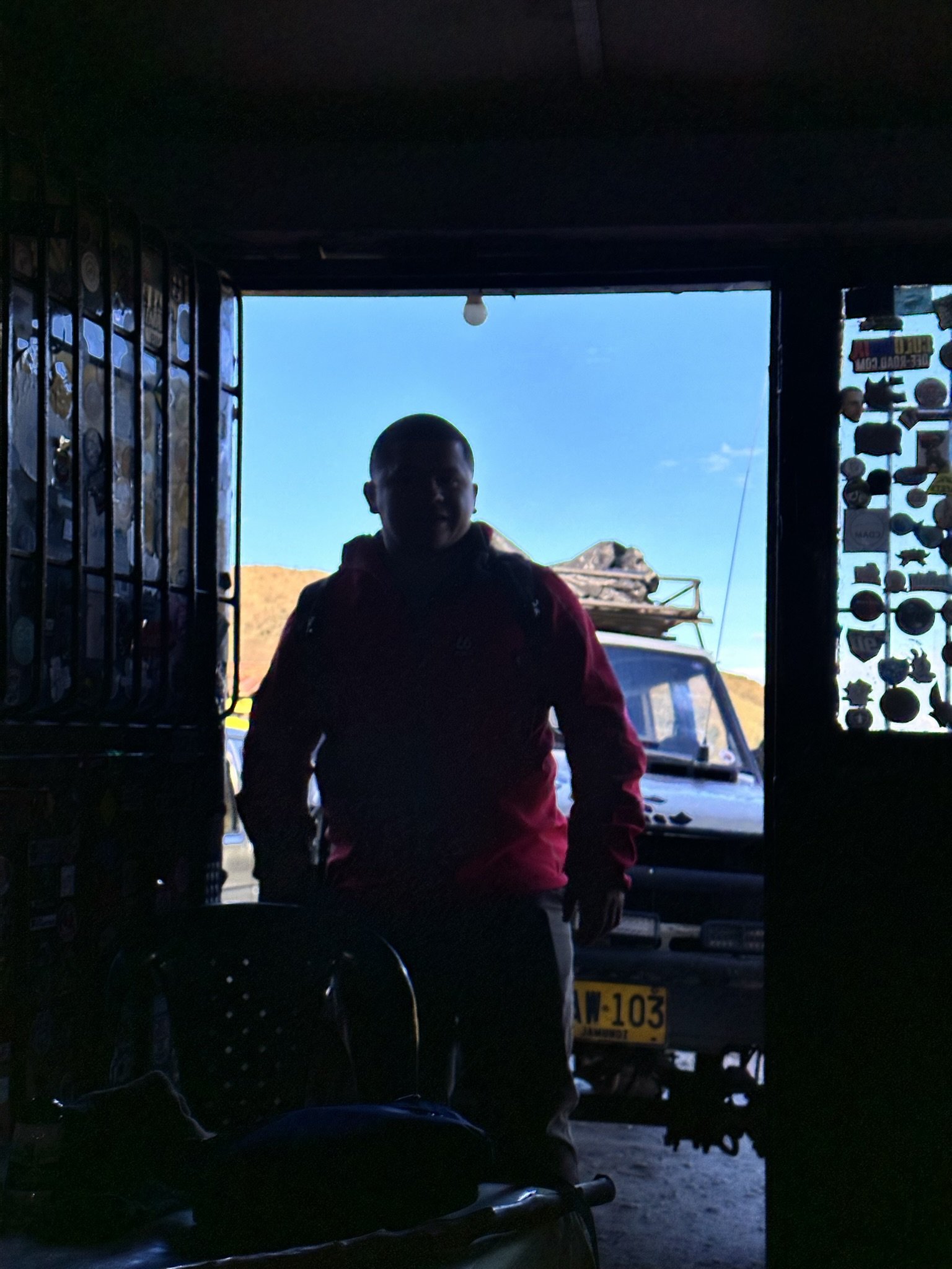 Person standing inside a shop, silhouette visible against bright blue sky outside, with vehicles parked outside.