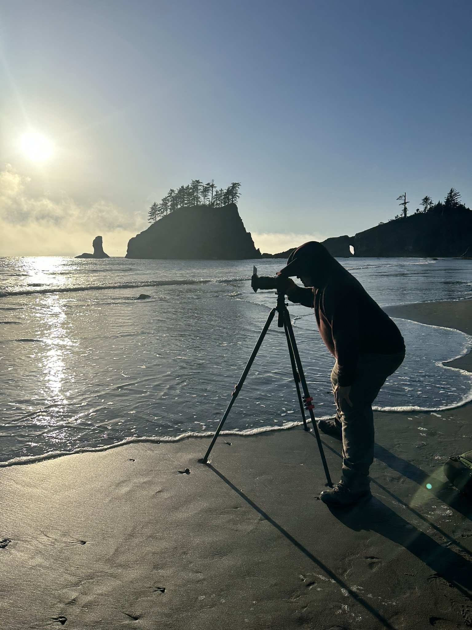 Person in hooded jacket taking photos with a camera on a tripod at the beach during sunset, with large rocks and trees in the background.