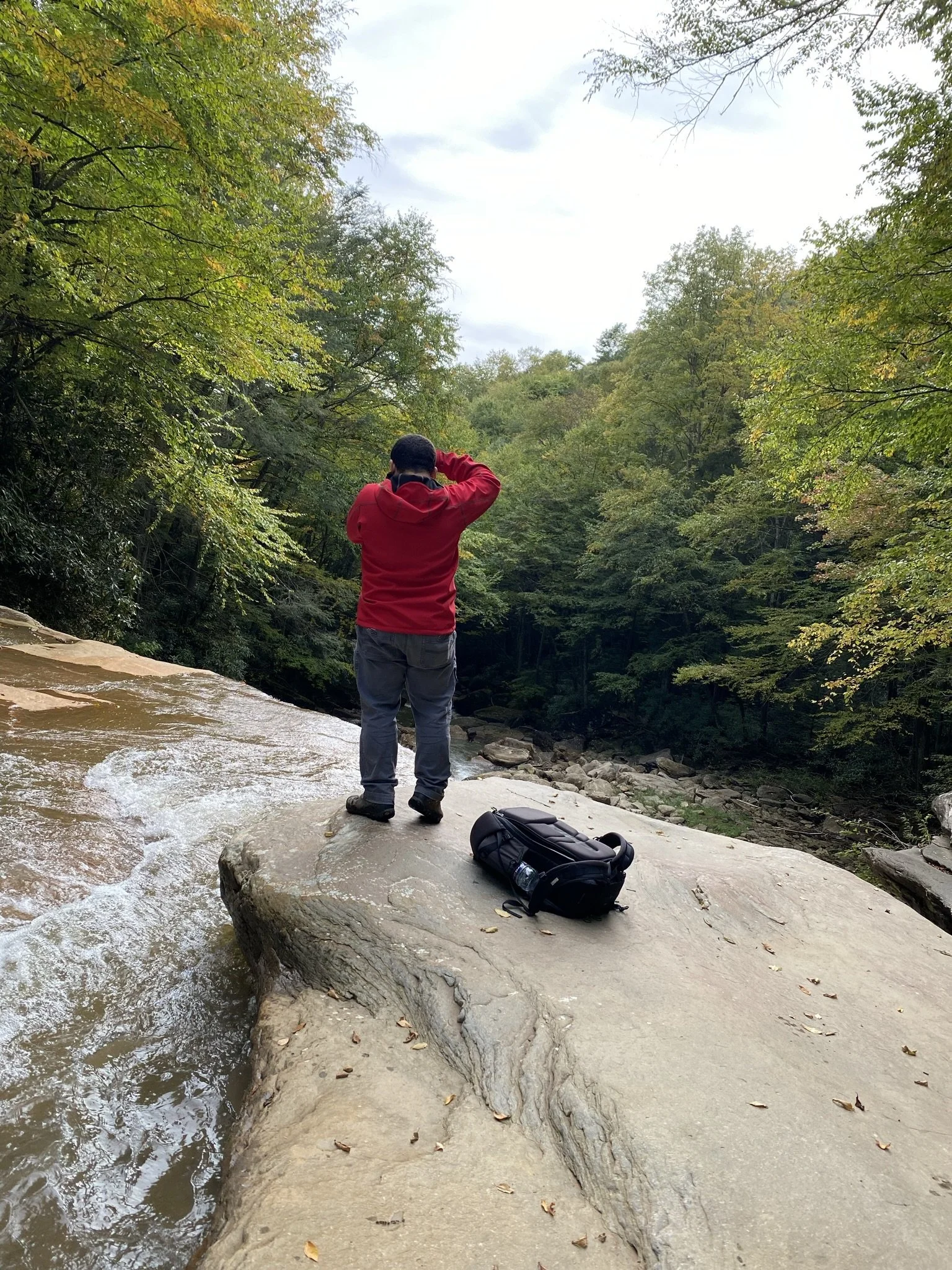 A person in a red hoodie standing on a large rock at the edge of a creek, holding their head with both hands, with a black backpack resting on the rock nearby. The scene is surrounded by green trees and an overcast sky.