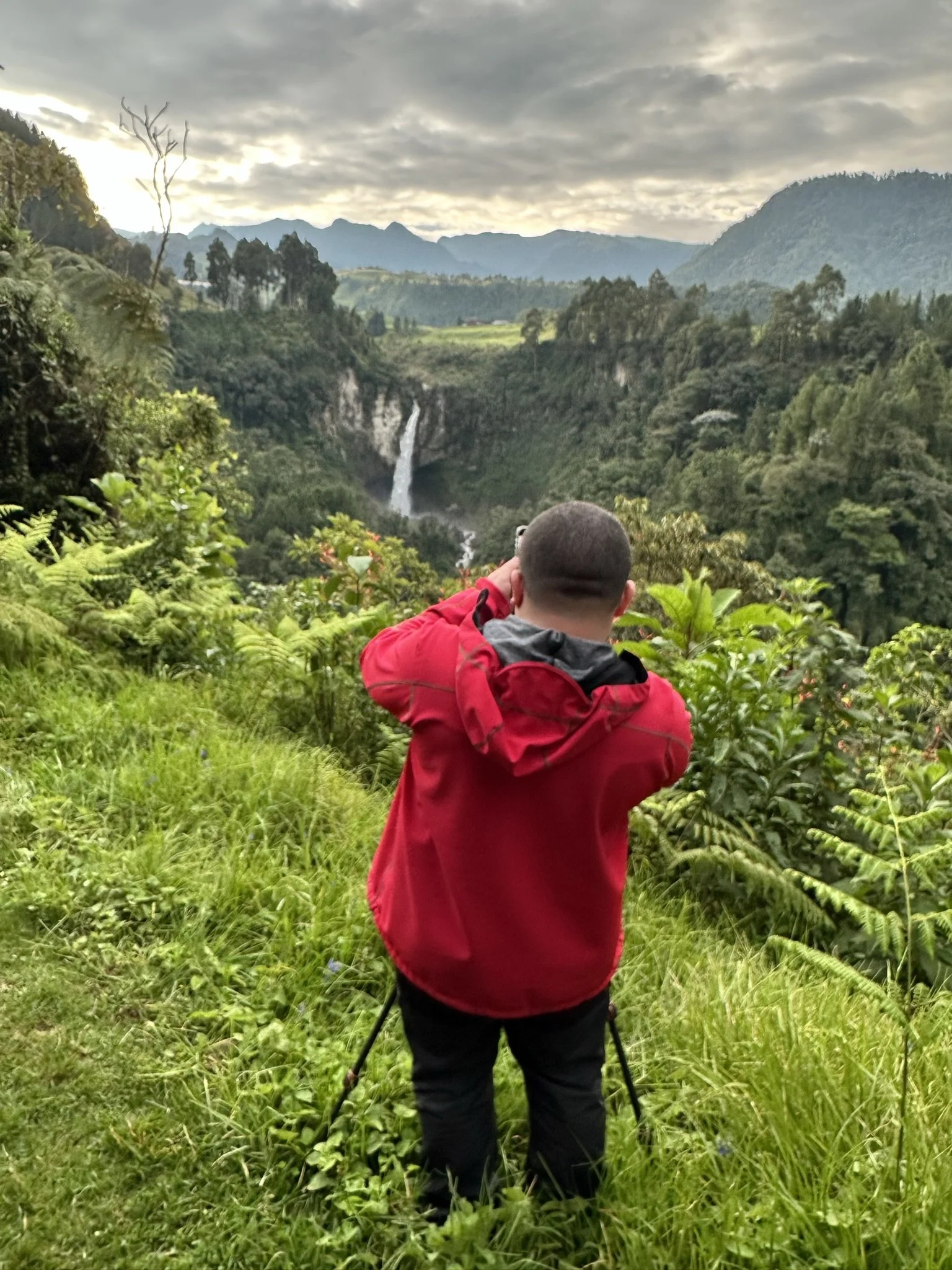 A person in a red jacket standing on a grassy hilltop, taking a photo of a waterfall in a lush green valley with mountains in the background.