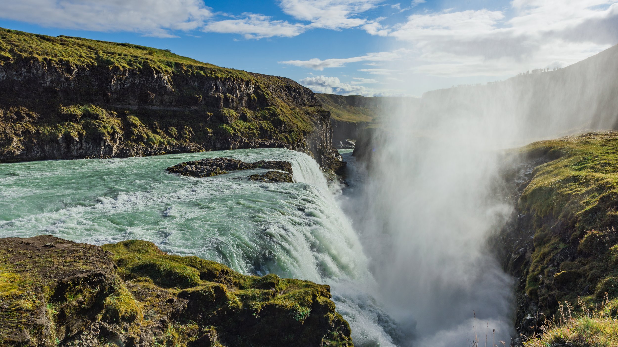 A scenic view of a waterfall flowing into a river below with green moss-covered rocks and grassy cliffs, under a partly cloudy sky.