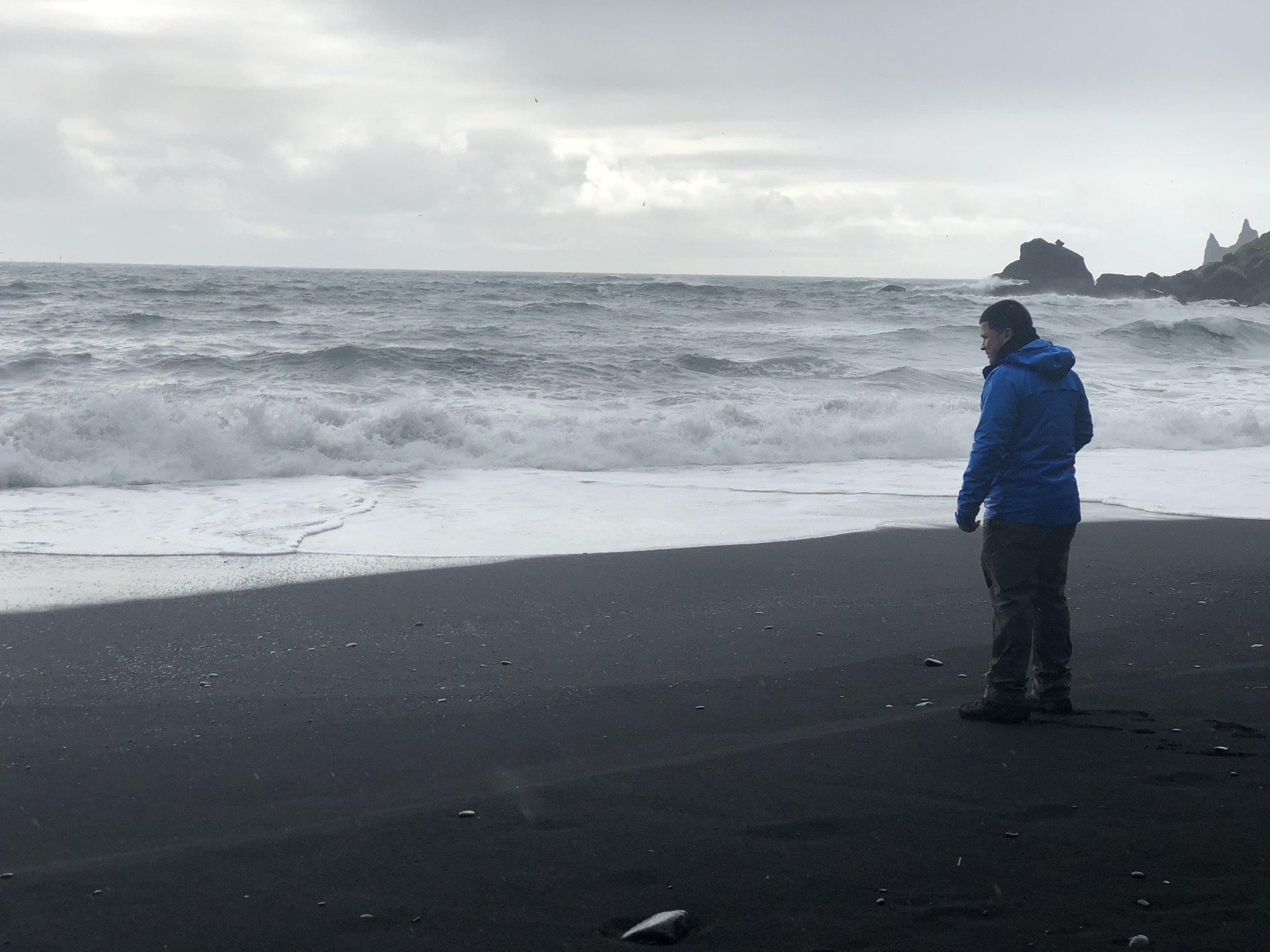 A person in a blue jacket stands on a black sand beach near the ocean, looking down at the wet sand with small rocks, with waves crashing nearby and rocks in the distance under a cloudy sky.