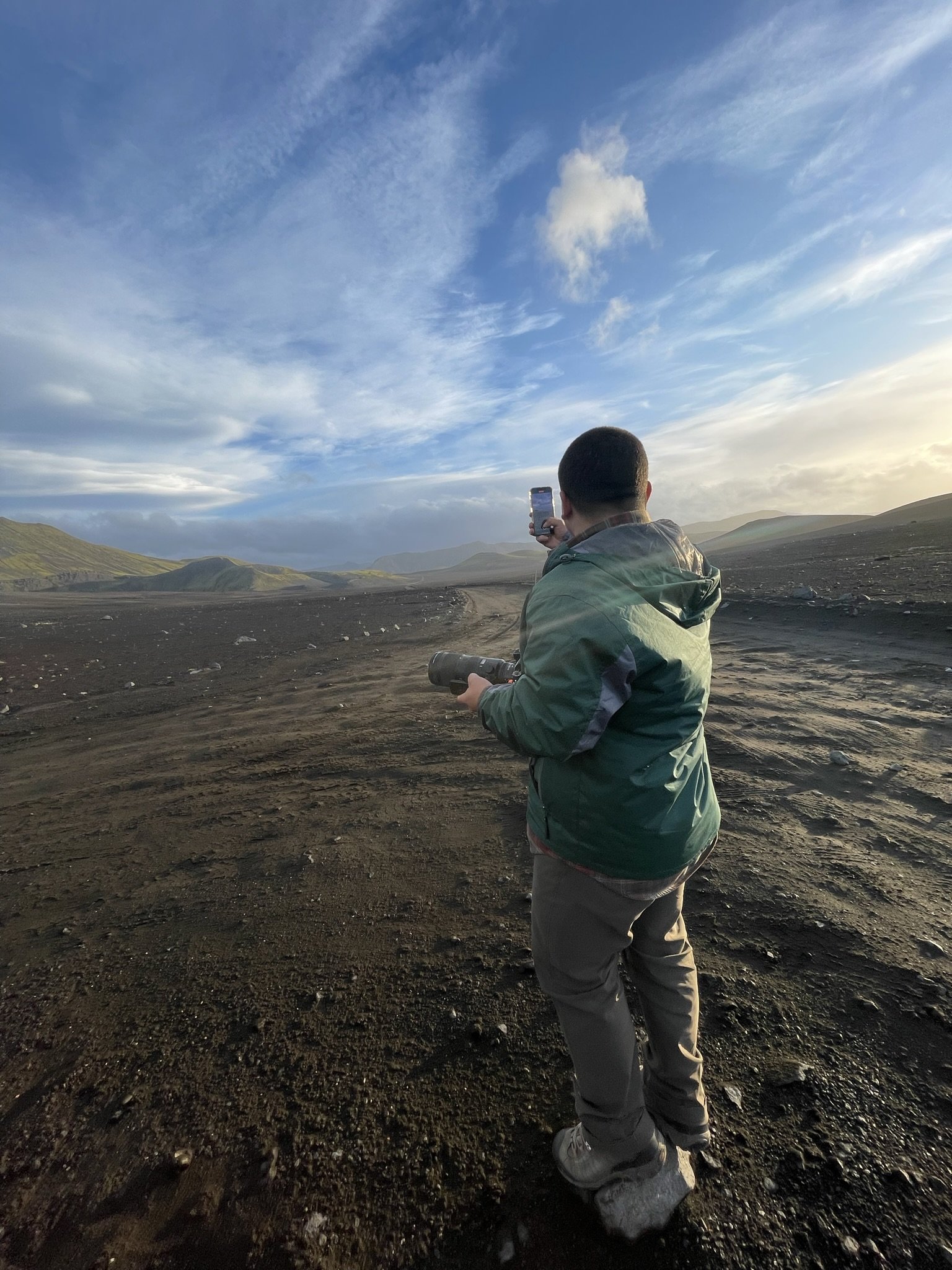A person standing on a dirt trail in a volcanic or barren landscape, taking a photo with a smartphone. The landscape includes distant hills and a partly cloudy sky.