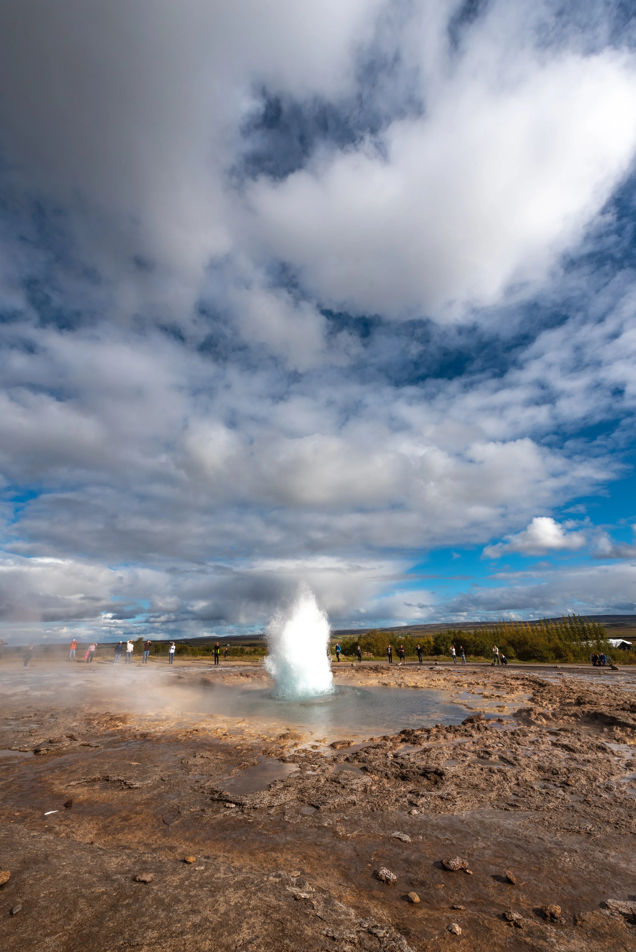 A geyser erupts with a tall fountain of water and steam in a geothermal area, with visitors walking and observing in the background under a partly cloudy sky.