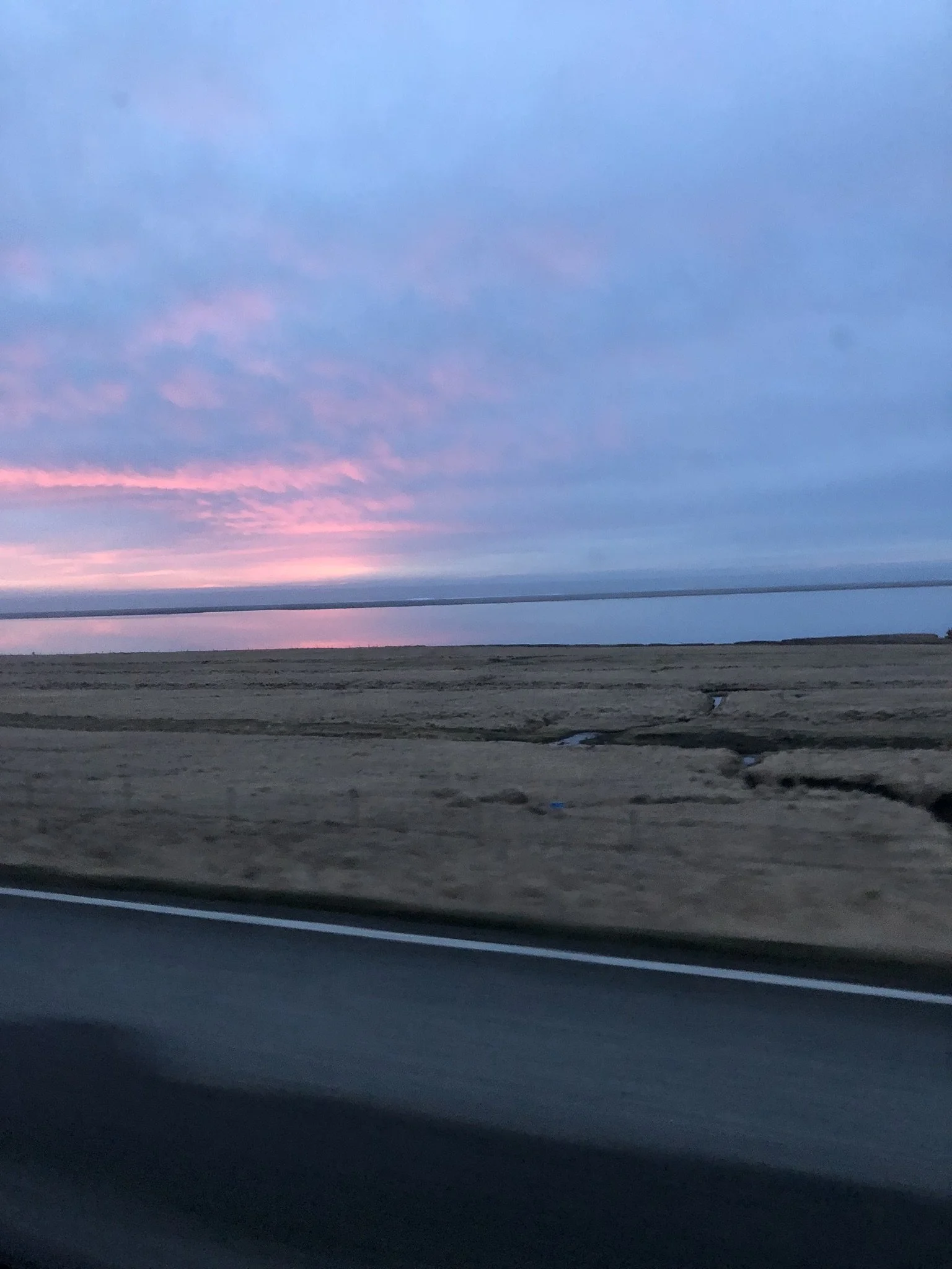 Photo taken from inside a vehicle showing an open field, a distant body of water, and a sky at sunset with pink and purple clouds.