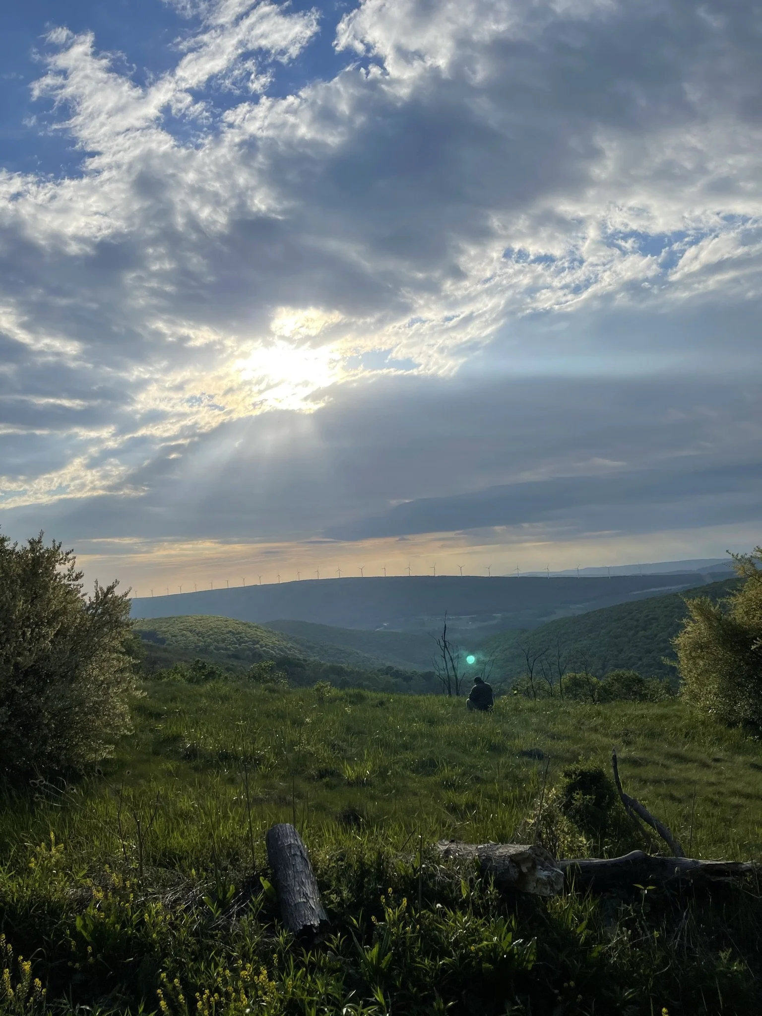 A person sitting in a green grassy field during sunset, with rolling hills, wind turbines on a distant ridge, and a partly cloudy sky with sun rays breaking through the clouds.