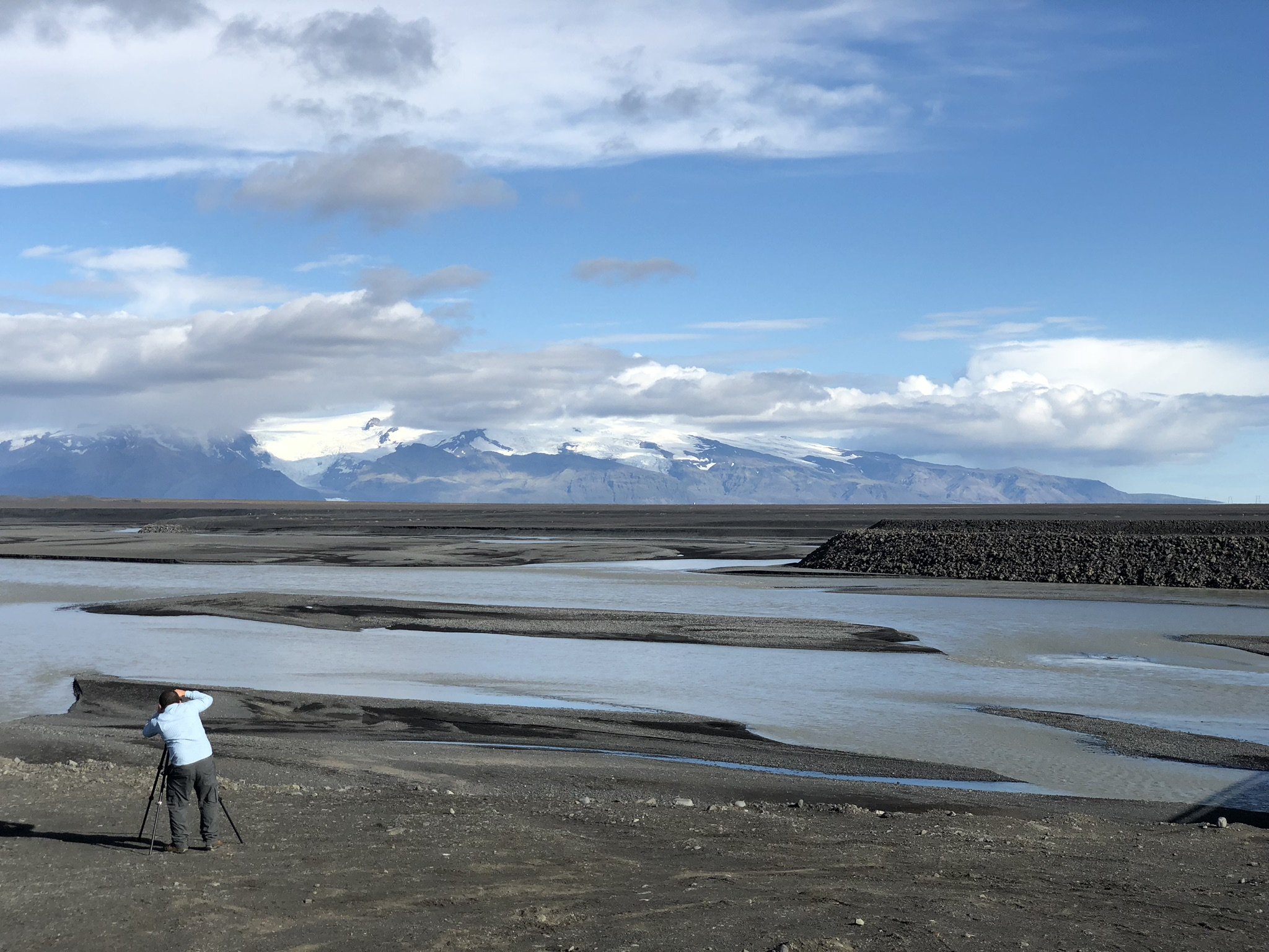 A man with a camera on a tripod standing on a gravelly shore, overlooking a river with snow-capped mountains in the background, under a partly cloudy sky.