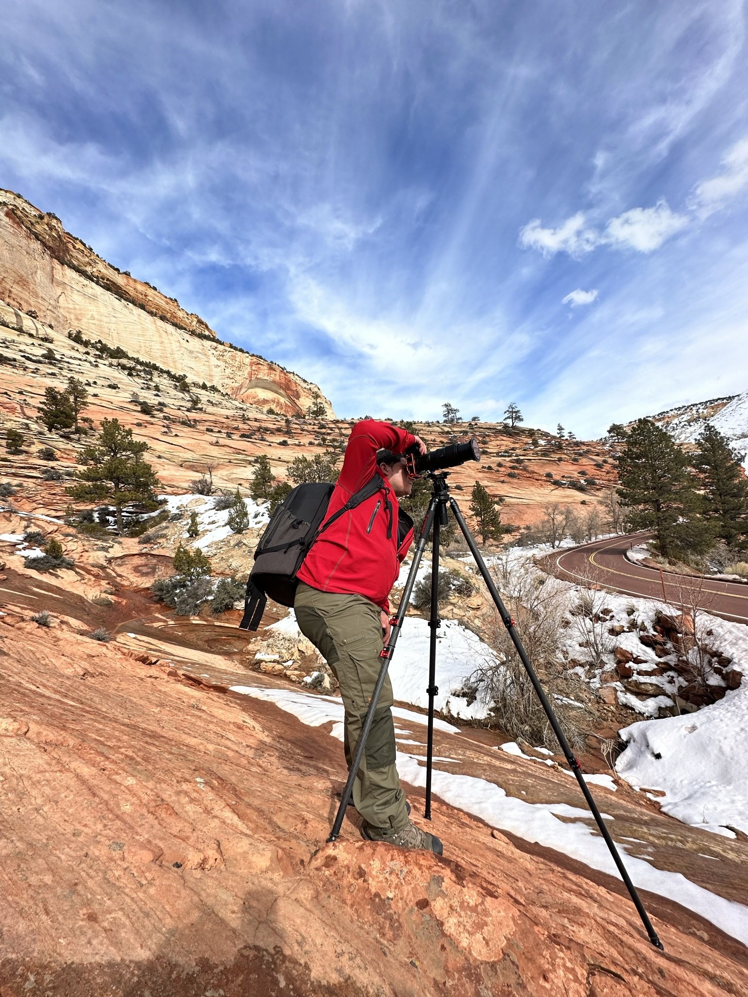 A person in a red jacket using a spotting scope on a tripod outdoors in a rocky, mountainous area with snow patches, trees, and a winding road beneath a blue sky with clouds.