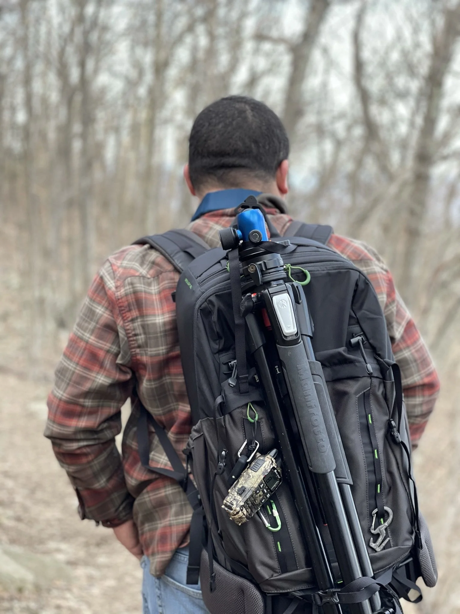Back view of a man in a plaid shirt carrying a large black backpack with a rifle and other gear attached, walking outdoors in a wooded area.