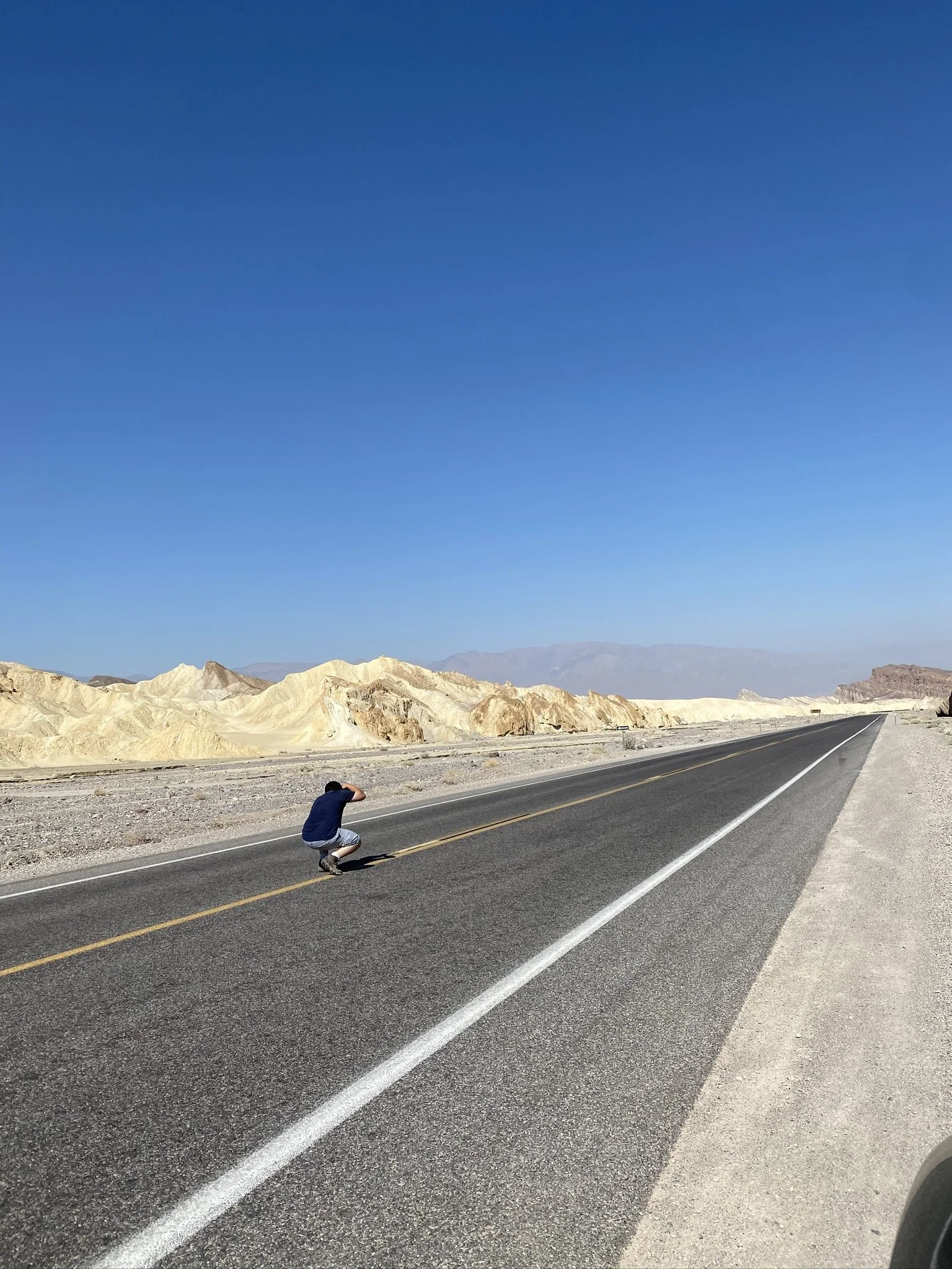Person crouching on the side of a long, straight desert road with rocky hills and mountains in the background under a clear blue sky.