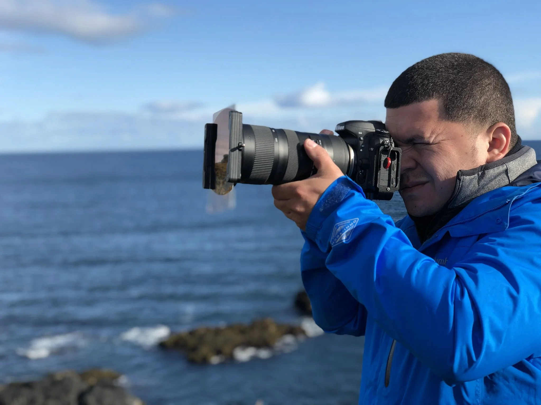 A man in a blue jacket takes a photograph with a professional camera near the ocean.