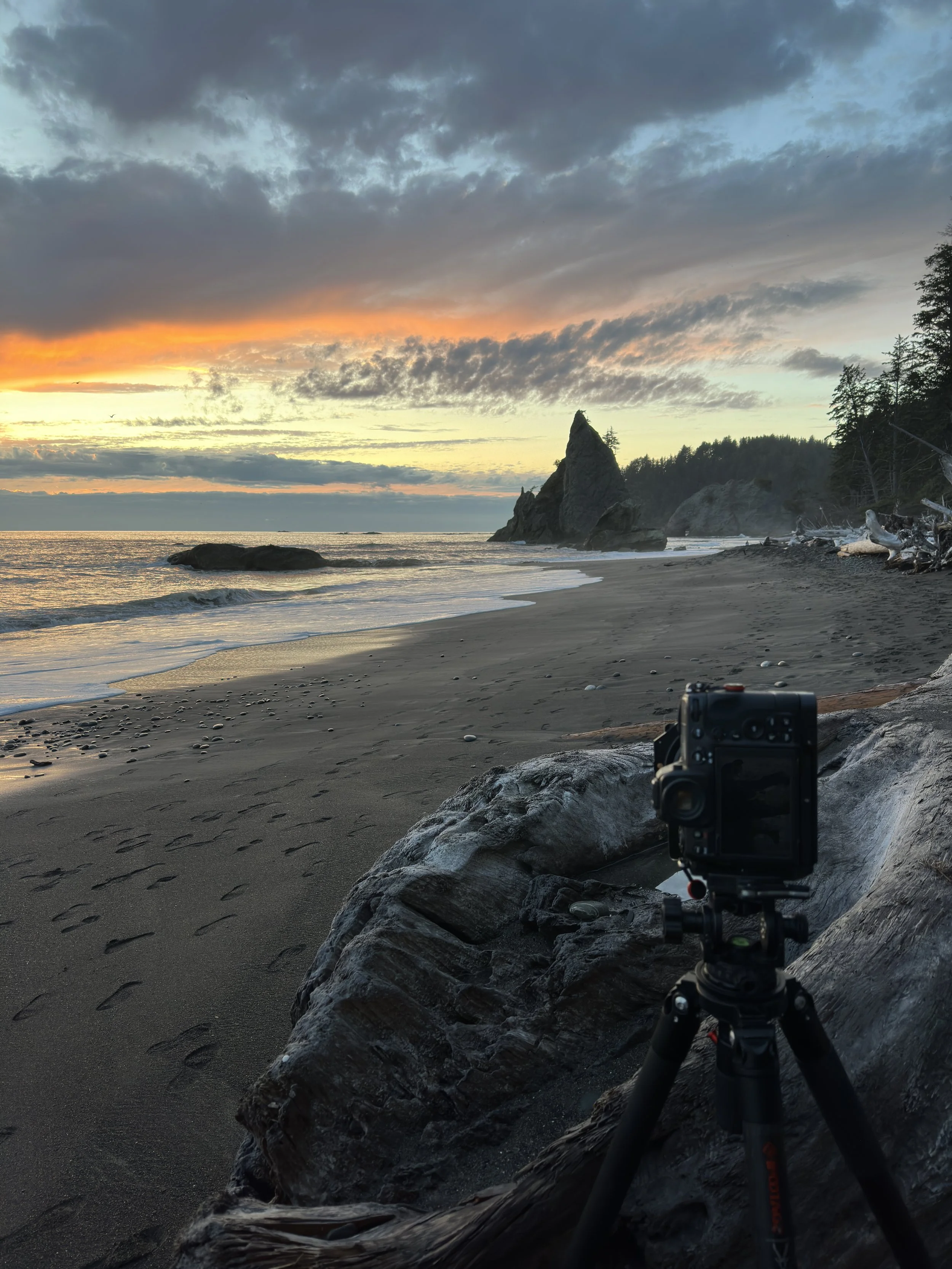 Camera set up on a tripod on a dark sandy beach capturing a sunset over the ocean with large rocks and driftwood, distant forested coastline, and dramatic clouds in the sky.