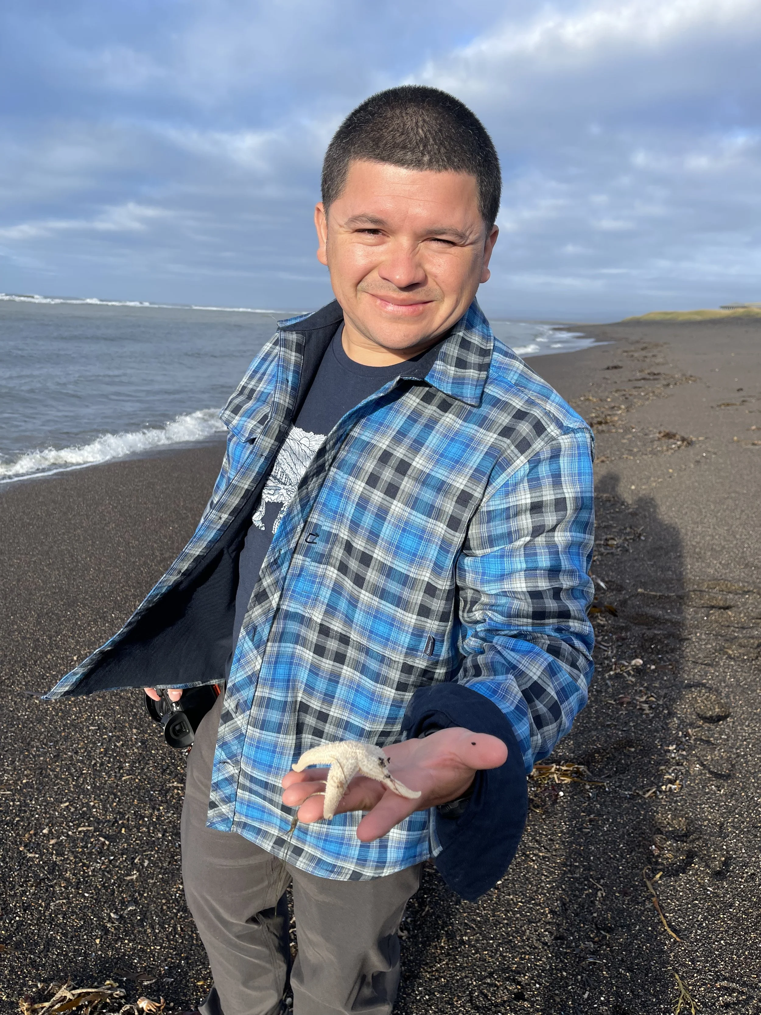 Melser holding a starfish on a beach with ocean waves and a cloudy sky in the background.