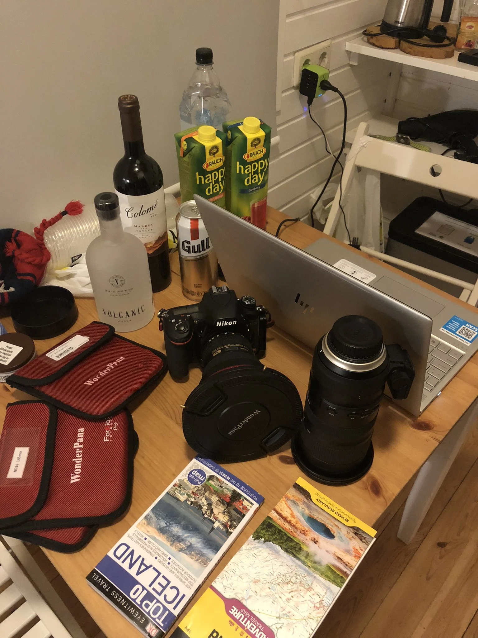 Cluttered wooden table with a Nikon camera, lens, red WonderPana cases, and travel brochures, surrounded by bottles of alcohol, juice, water, a laptop, and various electronic devices.