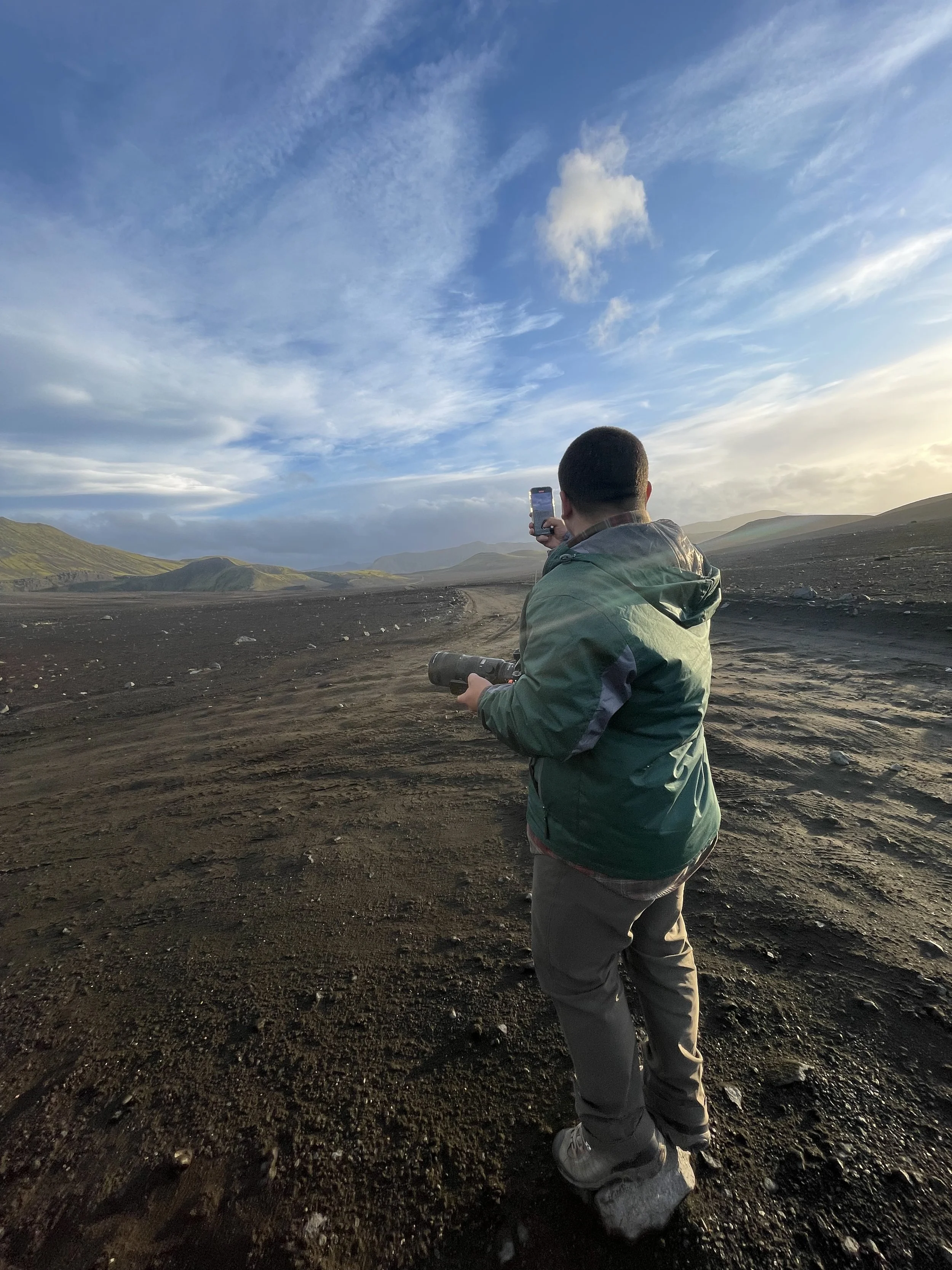 A person in a green jacket holding a camera and a phone, standing on a dirt road in a mountainous landscape with a blue sky and scattered clouds.