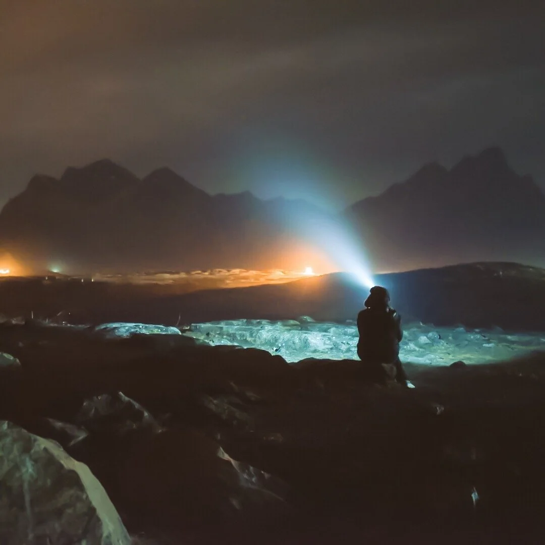 Person sitting on rocks near a coastline at night, illuminated by a flashlight, with mountains in the background and distant fires or lights on the horizon.