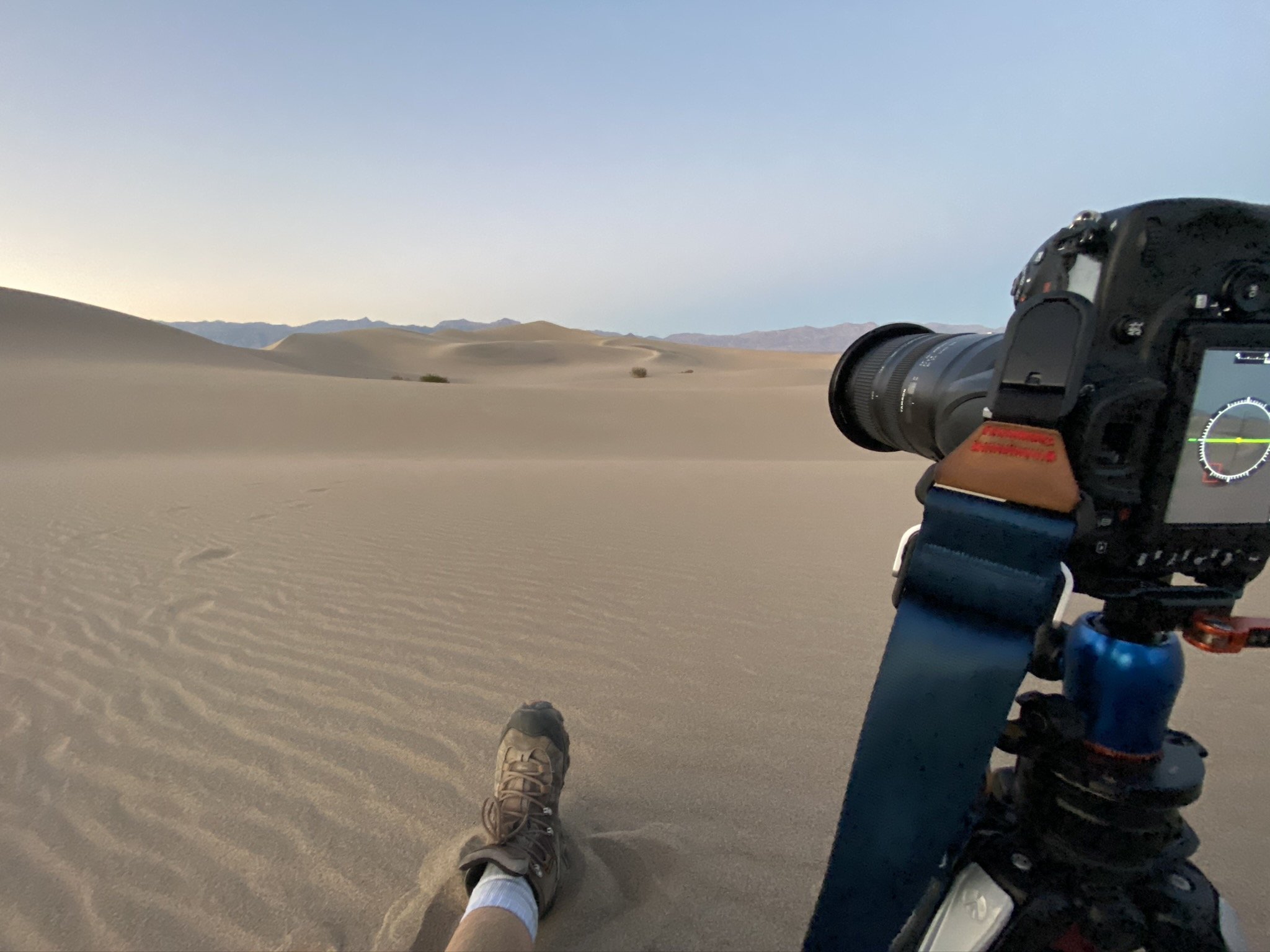 A person lying down on sand dunes with their foot and a camera mounted on a tripod visible, in a desert landscape with distant mountains under a clear sky.